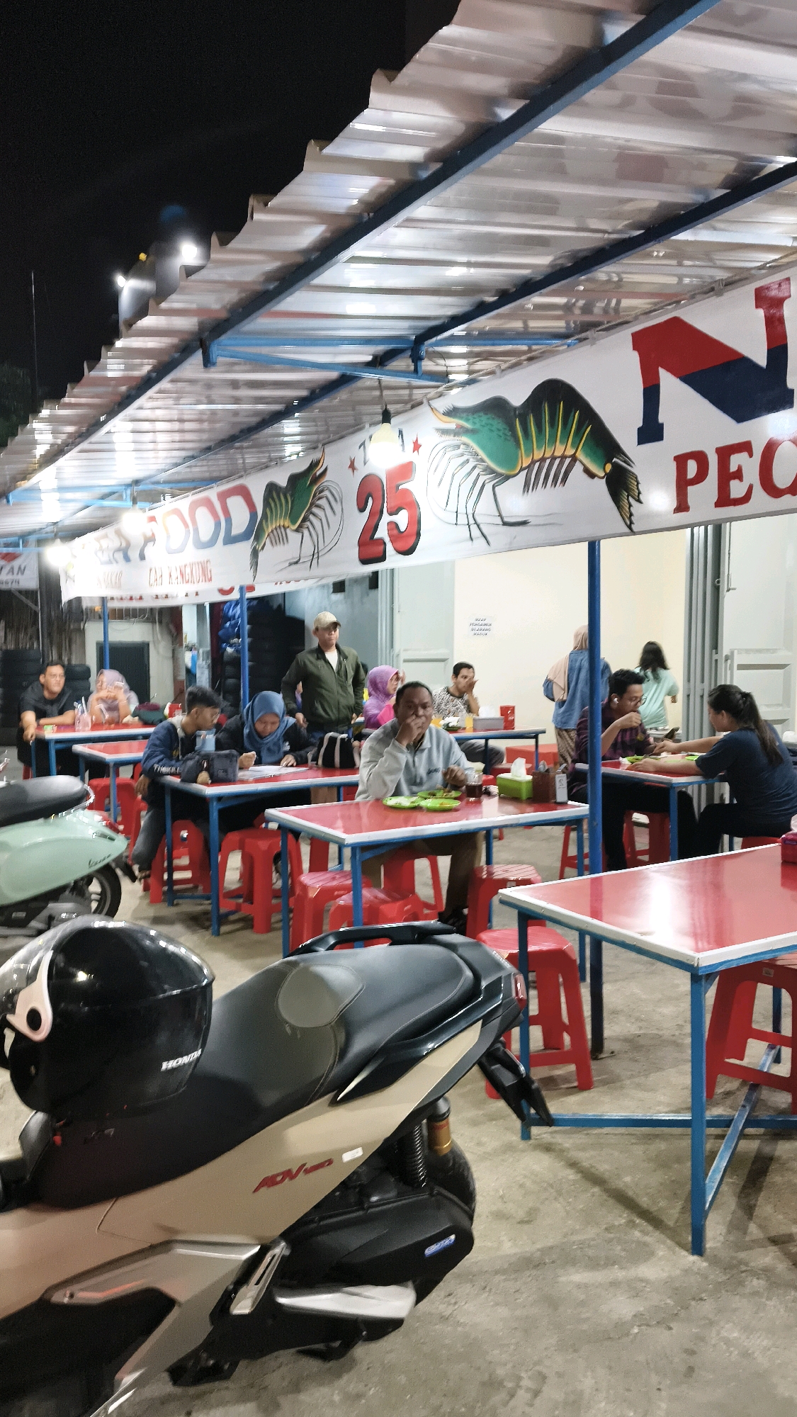 The image depicts an outdoor food stall, likely a seafood restaurant, situated in Karangsatria, Indonesia. The scene is illuminated by overhead lights and the ambient glow of the evening sky. A prominent banner above the seating area displays images of large shrimp and text, including "FOOD" and "PECO", suggesting a specialty in seafood. The restaurant features numerous tables with red tops and blue metal legs, surrounded by red plastic stools. Several people are seated at the tables, some eating, others engaged in conversation. A man in a beige cap and green jacket stands near the tables, possibly an employee. In the foreground, a motorcycle with a black helmet resting on its seat is parked, indicating a mode of transportation for patrons. The overall environment appears to be a casual dining setting.