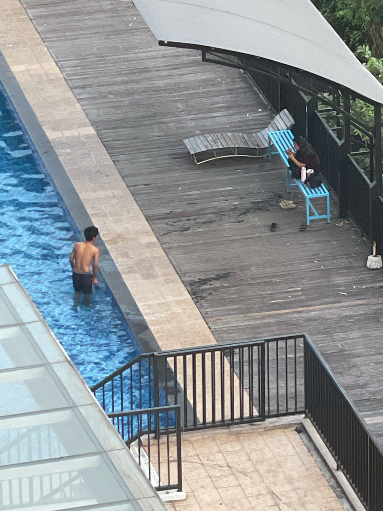 In an outdoor area, a young man stands in a swimming pool. The pool is rectangular and filled with blue water, with a tiled edge. Adjacent to the pool is a wooden deck. On the deck, a woman sits on a blue bench, holding a cup and looking down. A lounge chair is also visible on the deck. A black metal railing separates the deck from a tiled area. The scene is partially covered by a gray awning and surrounded by greenery.