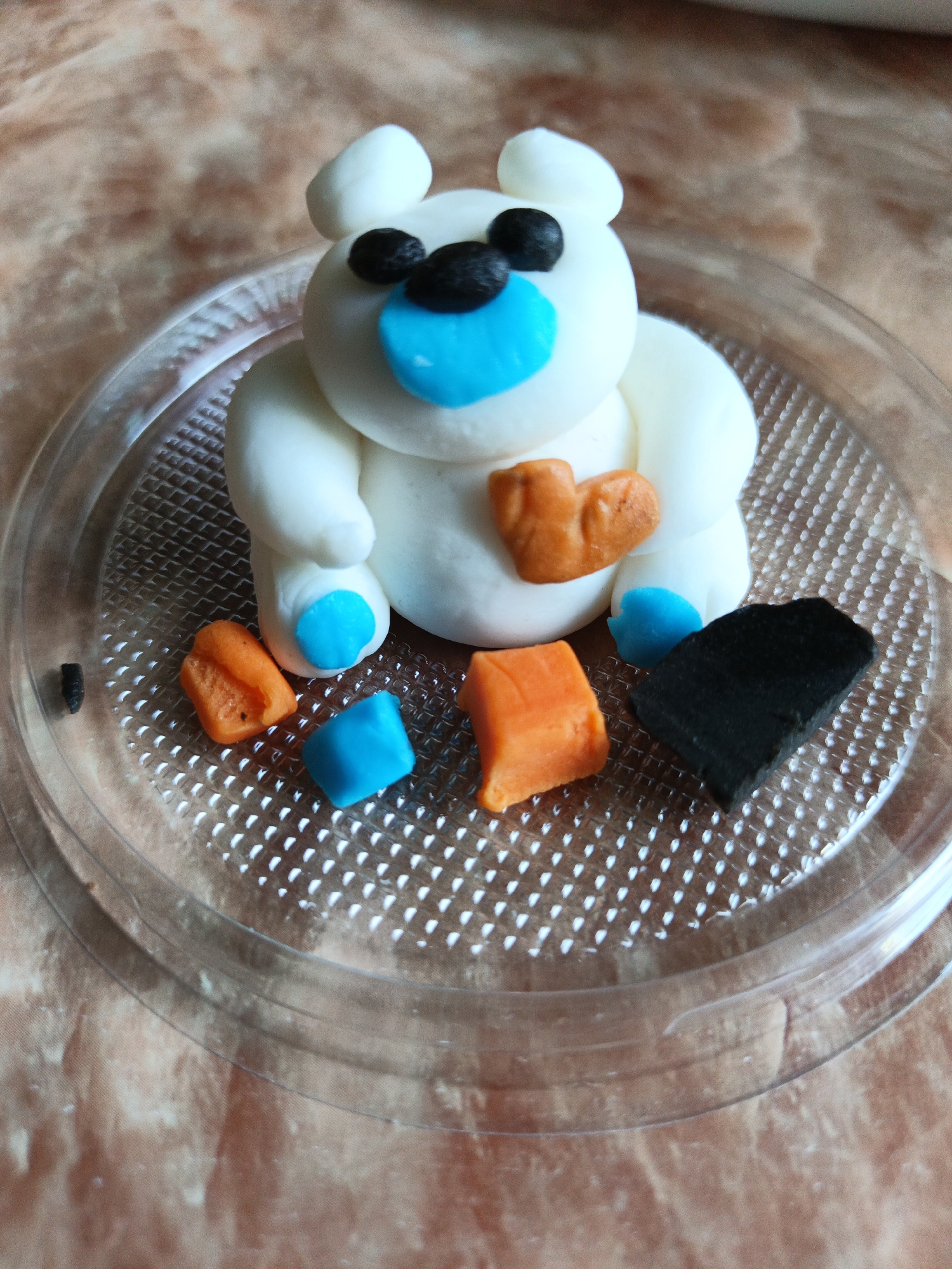 A handcrafted figure, resembling a white polar bear made of modeling clay, is centrally positioned on a clear, round plastic tray. The bear features a rounded white body, two black spherical eyes, a light blue heart-shaped nose, and two small white rounded ears. Its front paws are sculpted with a heart-shaped orange detail, and its feet exhibit light blue pads. Around the base of the bear on the tray are several scattered pieces of clay: two irregular orange pieces, one light blue cube, one black rectangular piece, and a small, dark cylindrical fragment. The clear plastic tray has a raised square grid texture. The entire arrangement rests on a blurry, light brown and off-white mottled surface, likely a tabletop or similar flat surface. This scene is located in the city of Лосева, Belarus.