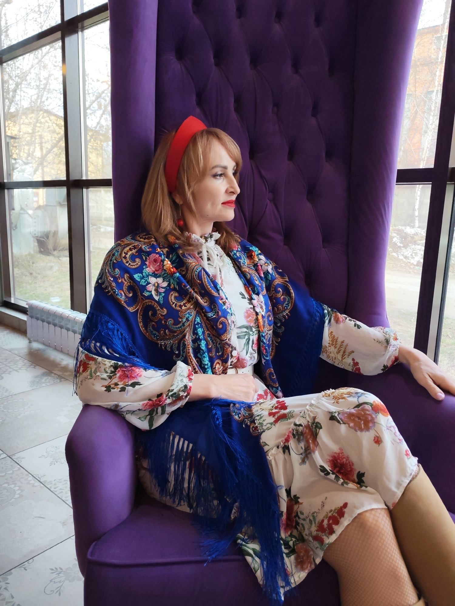 A woman with reddish-blonde hair and a red headband sits facing right in a large, high-backed purple armchair with tufted upholstery. She wears red lipstick and red dangle earrings. She is dressed in a white long-sleeved dress with a vibrant floral pattern in red, orange, and green hues, and fishnet stockings are visible on her legs. Draped over her shoulders and arms is a striking blue shawl featuring intricate gold, red, and green floral and paisley patterns, complete with a blue fringe. Her gaze is directed out of a large window to her right, conveying a calm and contemplative mood.
The setting appears to be an indoor space, possibly a café or waiting area, in Chaikovsky, Russia. The room features light-colored tiled flooring with a subtle design. Outside the expansive window, bare trees and distant buildings are visible, suggesting a cold season, likely winter or early spring. A white radiator is situated beneath the window on the left side of the frame. The scene is well-lit by natural daylight, indicating a daytime setting.