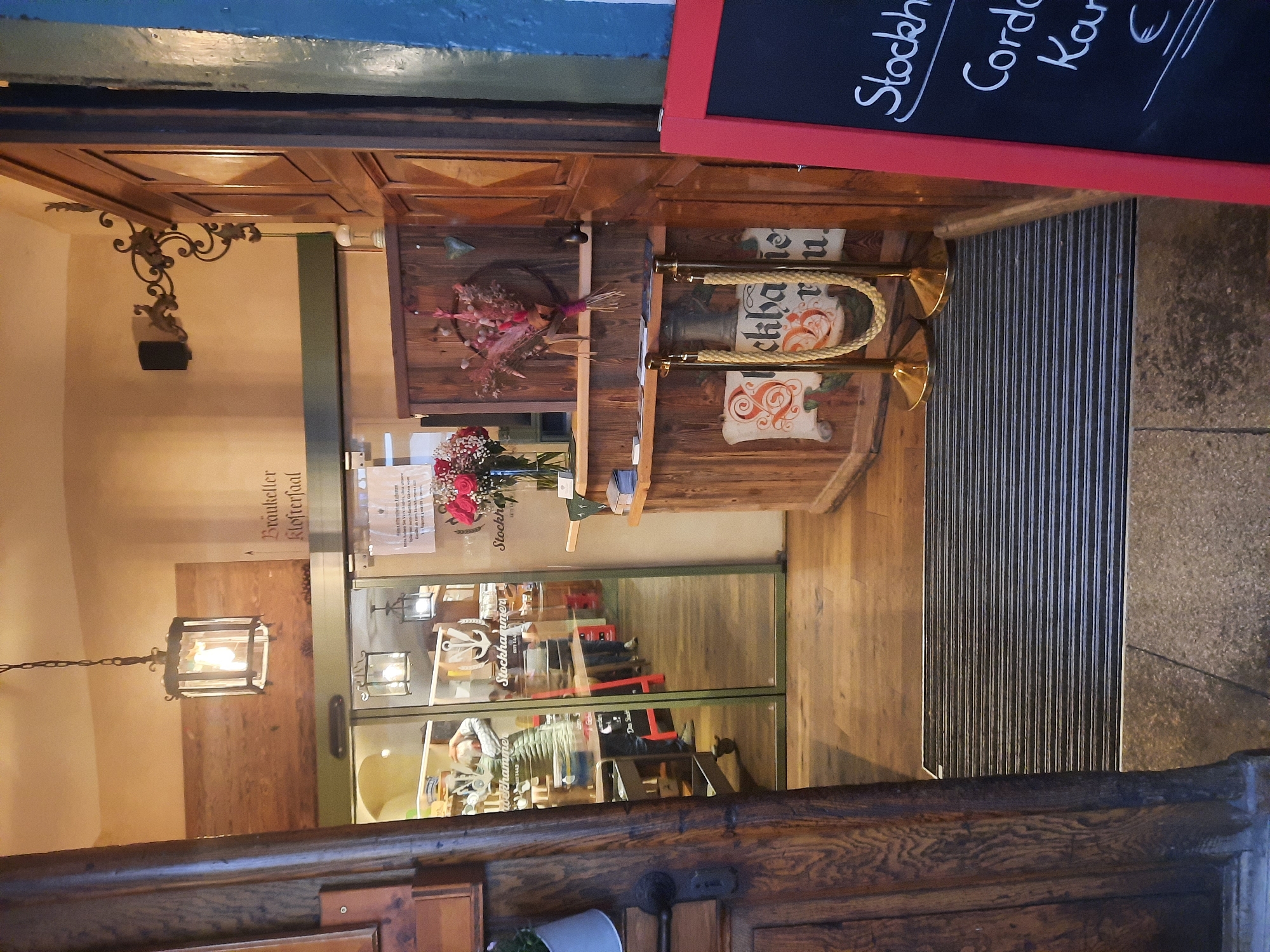 The image depicts the entrance to a establishment, likely a restaurant or pub, in Rosenheim, Germany. The facade is made of dark wood with glass doors that reflect the interior. Inside, the establishment appears to have a rustic, traditional ambiance with wooden decor. A person is visible through the glass doors, tending to the interior.
On the right side of the image, a wooden counter with a rope barrier suggests a reception or ordering area. A chalkboard menu with handwritten text is also visible. The overall scene conveys a welcoming and authentic atmosphere.
Based on the signs and signage visible in the image, the establishment appears to be called "Stockhamer" and is located in Rosenheim, Germany. The text "Bräukeller Kloster" also suggests a connection to a brewery or cellar.