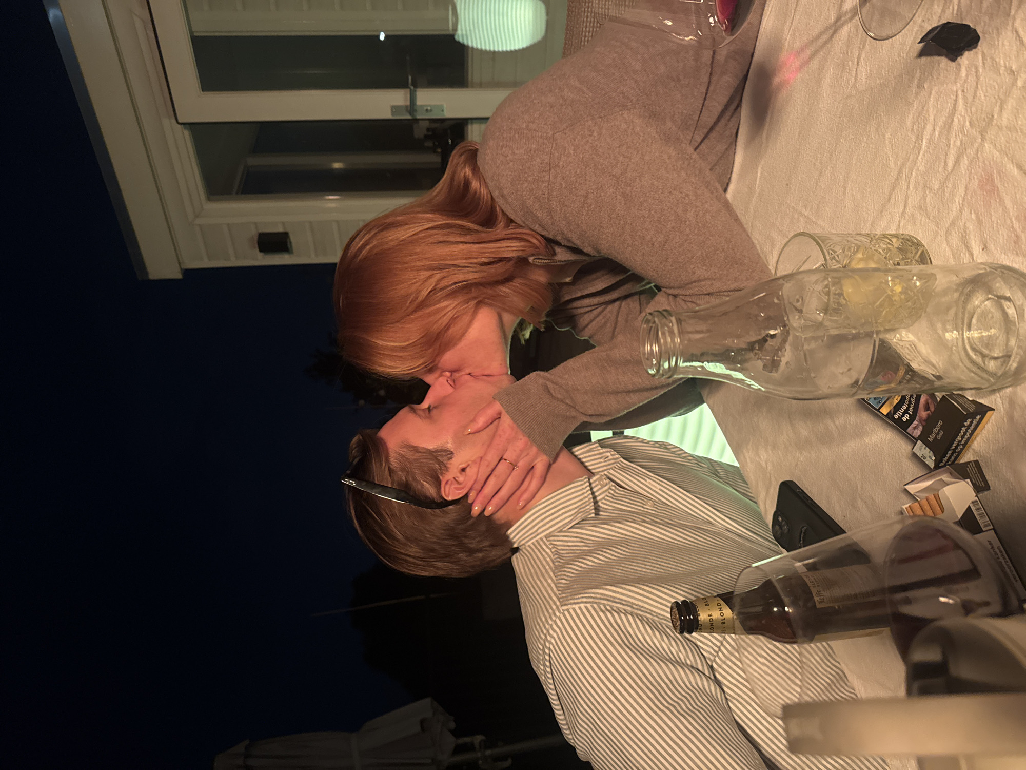 The image depicts a couple kissing at night, seemingly outdoors. A woman with red hair, wearing a gray sweater, is kissing a man in a striped shirt. The man is wearing sunglasses. They are seated at a table covered with a white cloth. On the table are glasses, a beer bottle, a bottle with a liquid inside, a phone, cigarettes, and a pack of Marlboro cigarettes with Dutch text. The background suggests an outdoor setting with a building visible and a dark sky. There is no verifiable information provided to identify Amsterdam, Netherlands.