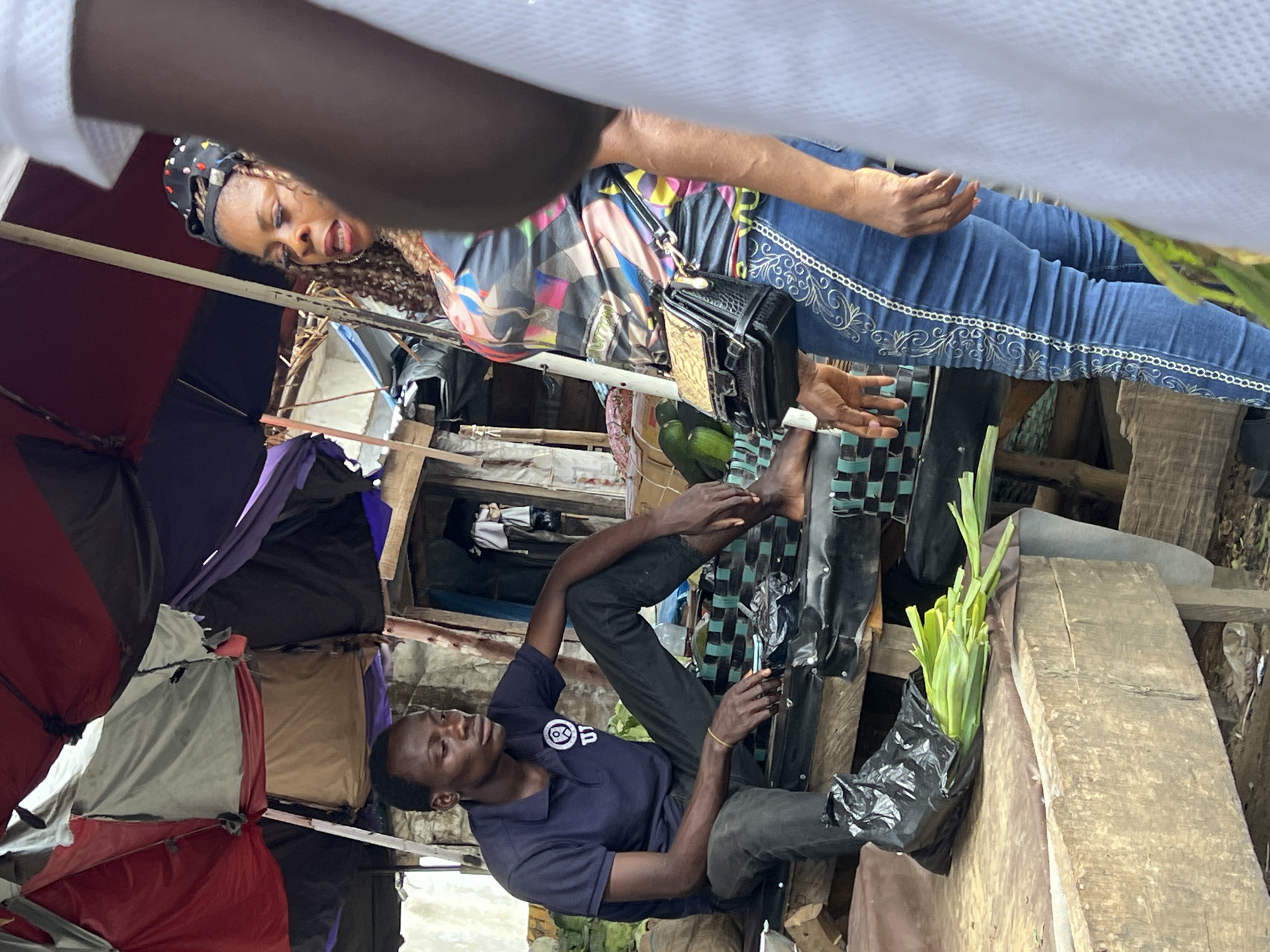 In Oko Amakom, Nigeria, a market scene unfolds under a colorful canopy of red, purple, and grey tarps. A young man in a dark blue polo shirt and black pants sits on a raised wooden platform, his legs crossed and arms resting on his knees. He appears to be looking down and to his right. Beside him, on the platform, is a black plastic bag containing a bunch of green leafy vegetables.
To the right of the young man, a woman stands with her body angled towards him. She wears a vibrant, multicolored top with a graphic print and dark blue jeans adorned with white embroidery along the outer seams of the legs. She has a black purse with a gold clasp slung over her shoulder. Her head is turned slightly, and she appears to be speaking. The background reveals more market stalls and structures, suggesting a bustling commercial environment. Another person, partially visible in white clothing, stands in the foreground on the far right, obscuring part of the woman. The ground beneath the platform is dusty and appears to be a mix of dirt and debris.