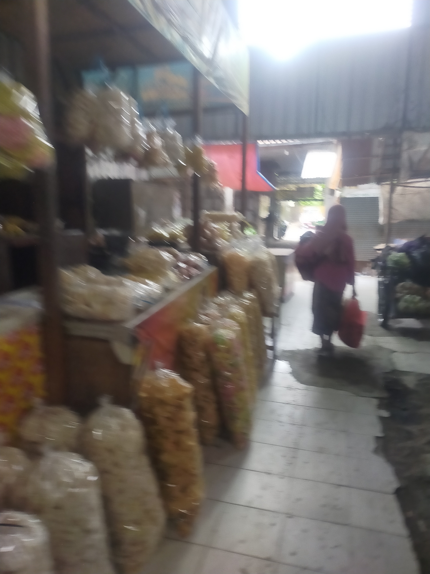 The image depicts a market scene in Trayeman, Indonesia, with an aisle of food stalls on the left and a person walking away from the camera on the right. The stalls are filled with packaged food items, primarily snacks in clear plastic bags. The floor is tiled and appears wet in places. The person on the right is wearing a pink headscarf and a dark skirt, carrying two bags, one red and one dark-colored. The lighting suggests it is either early morning or late evening. The overall atmosphere is that of a bustling local market.