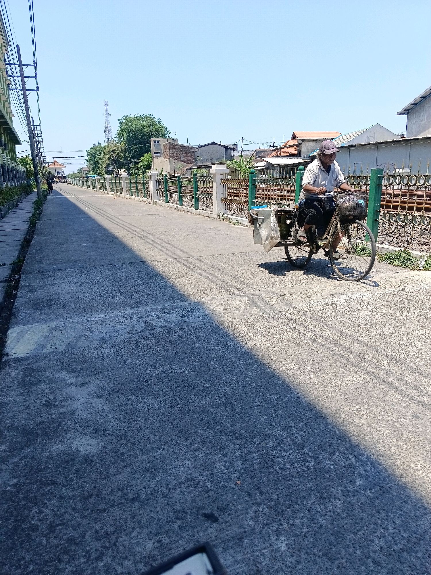 In the city of Kalijaten, Indonesia, on a clear, sunny day, an elderly man with a light-colored shirt, dark pants, and a cap rides a bicycle on a paved road. The bicycle has a basket on the front containing items and a larger carrier with bags attached to the rear. The man is positioned in the right half of the frame, moving away from the viewer. The road is mostly empty, with a shadow cast across it from the left, indicating the sun's position.
To the left of the road, a sidewalk is visible, lined with a building with green and white accents and electrical poles with wires. Further down the road, another person walks away from the camera.
To the right of the road, a green metal fence with decorative patterns separates the road from a train track area. Behind the fence, various buildings with different roof styles and colors, along with some trees, are visible. A communication tower is also present in the distance, beyond the buildings. The overall environment suggests a suburban or semi-urban setting.