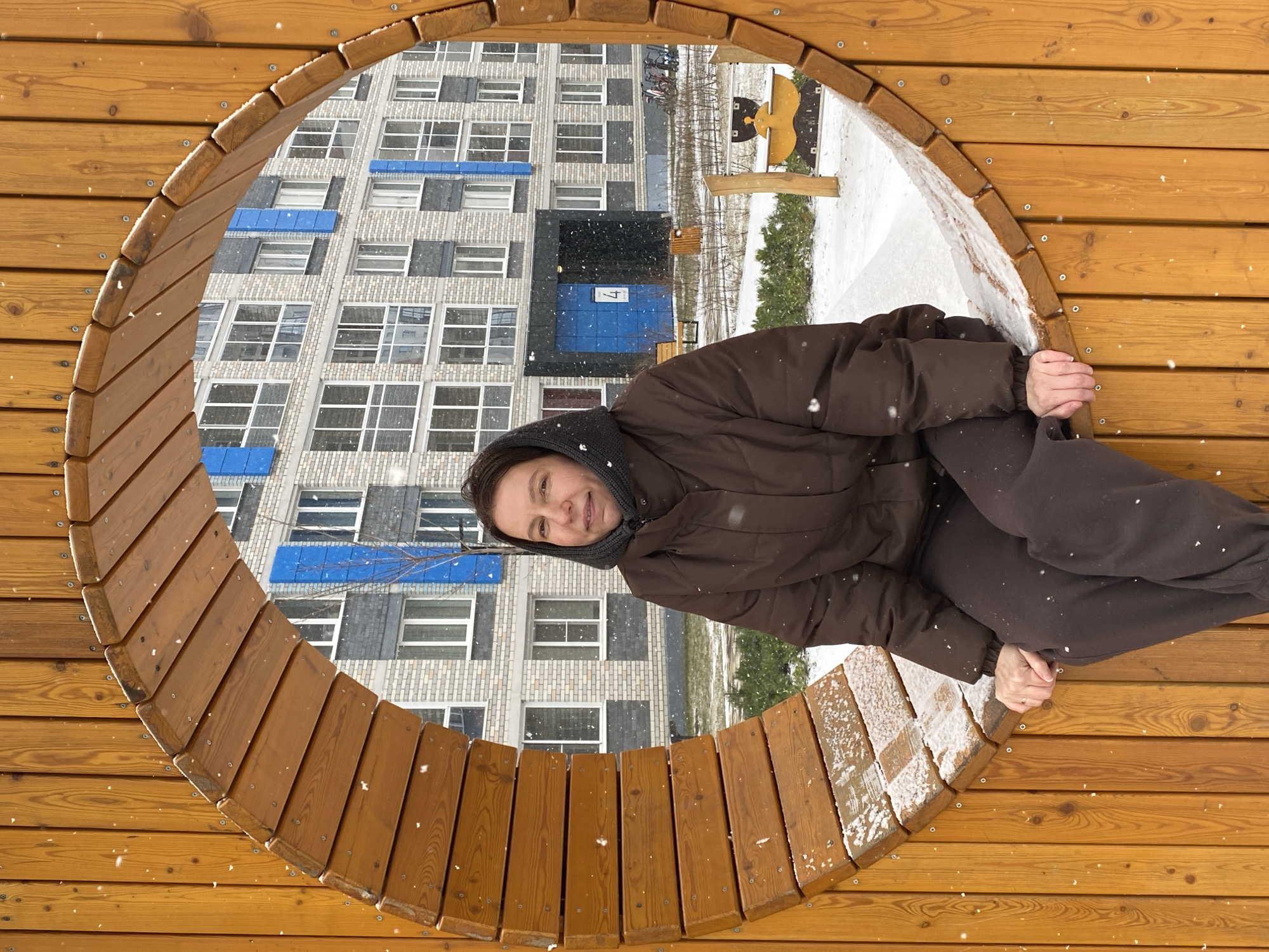 A woman in a brown jacket and hood is seated inside a circular wooden structure with snow. The backdrop features an apartment building with a blue entrance and white-framed windows, indicating the location is Yekaterinburg, Russia. Snowflakes are visible in the air, suggesting a winter setting.