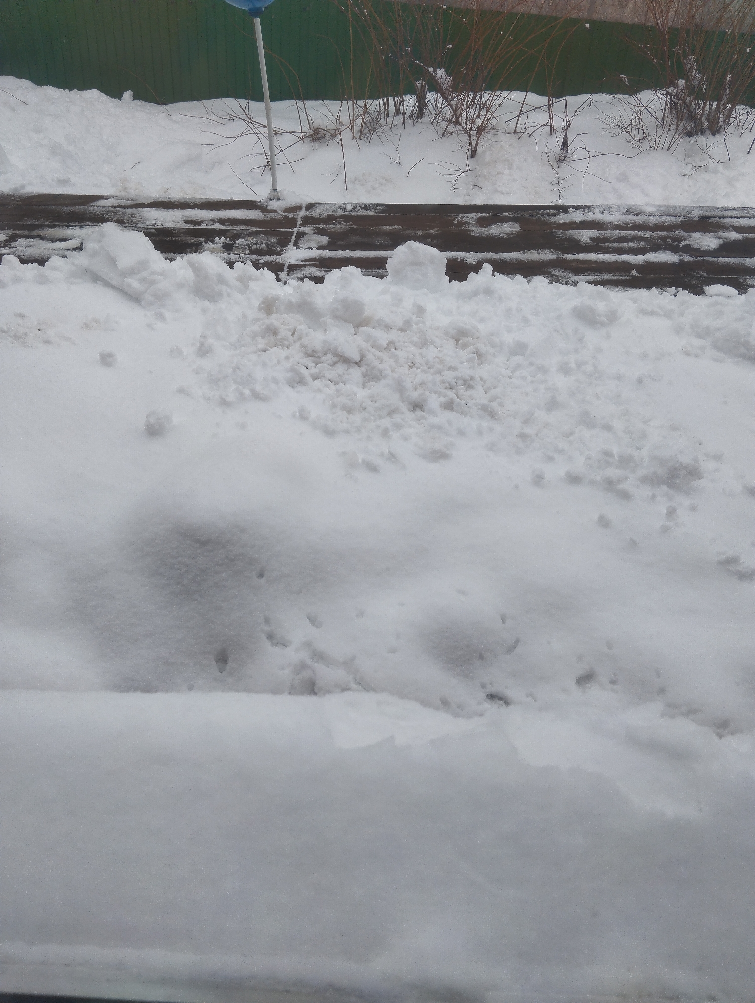 The image depicts a snow-covered scene in what appears to be an outdoor location. In the background, there's a green fence or wall. In front of the fence is a brown wooden structure, partially obscured by snow. The majority of the image is dominated by various textures of snow, suggesting it may have been recently moved or plowed. The photo was taken in the city of Krasnogorskoye, Russia. No people or animals are visible in the image. A blue sign is present. Brown vegetation is observed, and is mainly concentrated in the upper right.