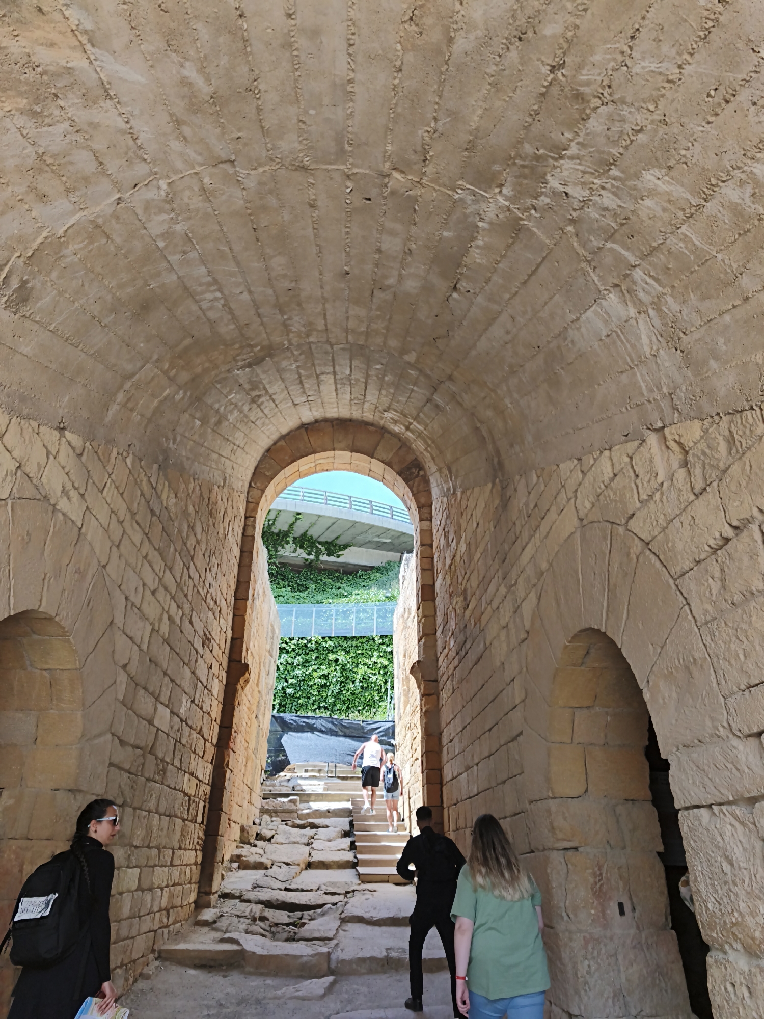 Here's a detailed description of the image:
**Scene:** The image depicts the interior of a stone archway or tunnel, likely part of an ancient structure. The tunnel has arched ceilings and walls constructed from rough-hewn stone blocks. Light enters from the opposite end of the tunnel, where steps lead upwards toward an opening revealing a bridge-like structure and greenery.
**People:** Several people are visible within the scene.
* A woman in a black outfit and a backpack stands in the foreground on the left side of the tunnel.
* A person in a black outfit is walking towards the steps in the center.
* A woman with long hair is walking towards the stairs wearing a green shirt and blue jeans.
* In the distance, near the opening, are two people walking up the steps.
**Objects:**
* Stone archway/tunnel structure
* Stone steps
* Backpack
* Bridge-like structure
* Green vegetation
**Environment:** The setting appears to be outdoors, with sunlight illuminating the end of the tunnel. The presence of old stones and the arched architecture suggests a historical site.
**Location:** The location is Tarragona, Spain.