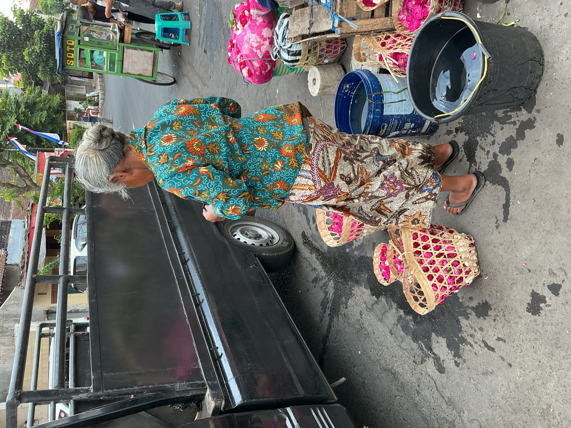 A rear view shows an elderly woman with grey hair in a bun wearing a colorful batik shirt and skirt standing next to a black truck. She is on a wet, paved street in Magelang, Indonesia. Around her are several woven baskets filled with pink and white flower petals. To her right are two buckets, one blue and one black, filled with water, and a green food cart with "SOTO" written on it. In the background, other people are visible near the food cart, and buildings line the street. The scene appears to be a market or roadside stall.