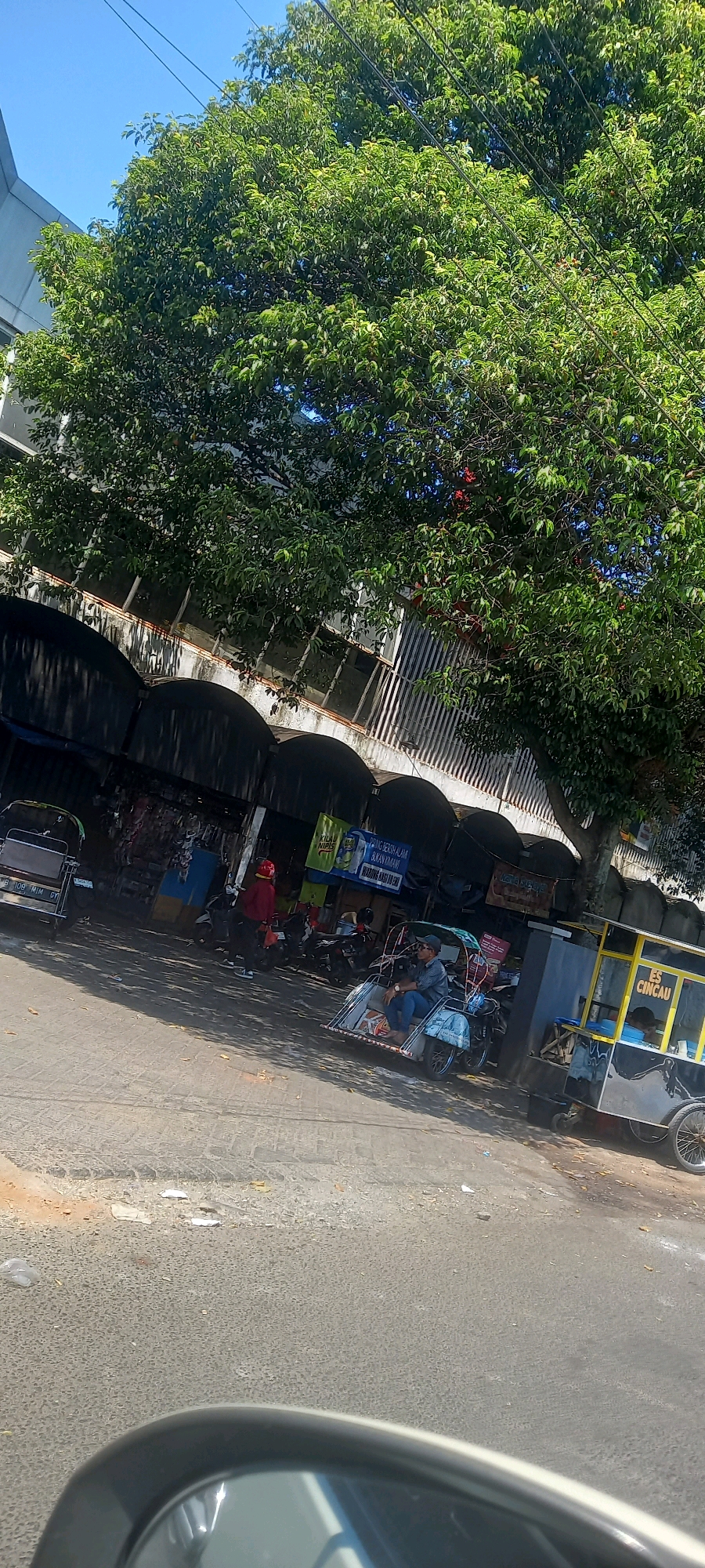 The image depicts a street scene in Jember, Indonesia, under a clear blue sky. A large, leafy tree dominates the upper portion of the frame, its branches casting shadows on the pavement below. In the background, a long building with arched openings runs along the street. In front of the building, there are several market stalls with tarps and wares displayed.
Several people are present. A person in a red jacket and red helmet is standing near the stalls. Another person, wearing a blue shirt and jeans, is seated in a three-wheeled vehicle that resembles a pedicab. This pedicab has a canopy and appears to be stationary. Further to the right, a food cart is visible. The cart is yellow and black and has the words "ES CINCAU" (Grass Jelly Drink) written on it. A person appears to be working at the cart.
On the street, there are a few motorcycles parked. Another three-wheeled vehicle, similar to the pedicab, is parked on the left side of the frame. The pavement shows signs of wear and tear, with some debris scattered on it. A car's side mirror is visible in the lower foreground, suggesting the photo was taken from a vehicle. The overall scene suggests a bustling market area or roadside commerce.