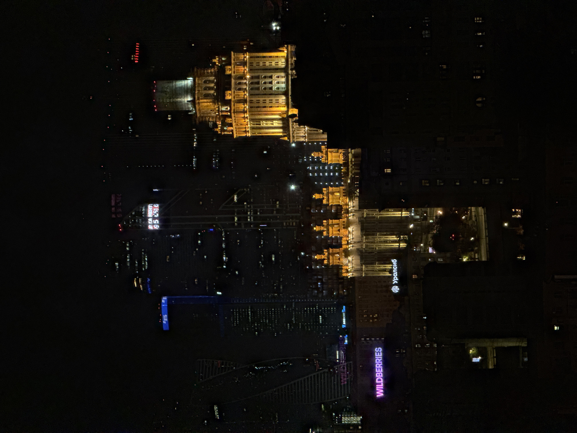Here's a description of the image:
The image shows a nighttime cityscape in Khimki, Russia, taken from an elevated perspective. Prominent features include brightly lit buildings and towers, notably the State University building with elaborate illuminated facades. Advertisements for "Wildberries" and "Uralsib" are also visible on buildings. The image shows modern buildings and skyscrapers, also reflecting the city's recent urban development.