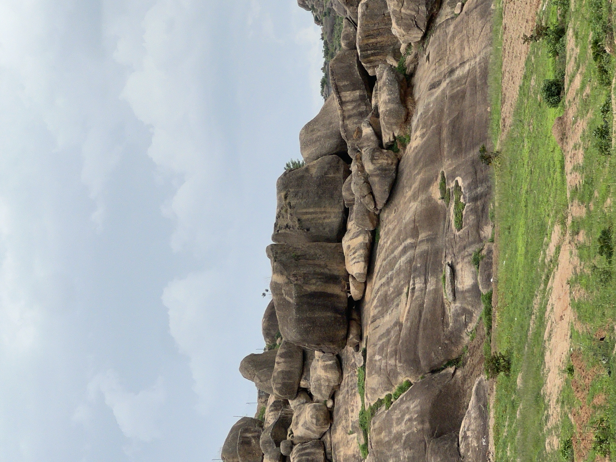 A medium shot captures a rocky, mountainous landscape under a partly cloudy sky. The rocks are layered and appear to be gray or brown, and they are partially covered in grass and vegetation. A patch of green grass is visible on the right side of the frame. The location is near the city of Jos, Nigeria.