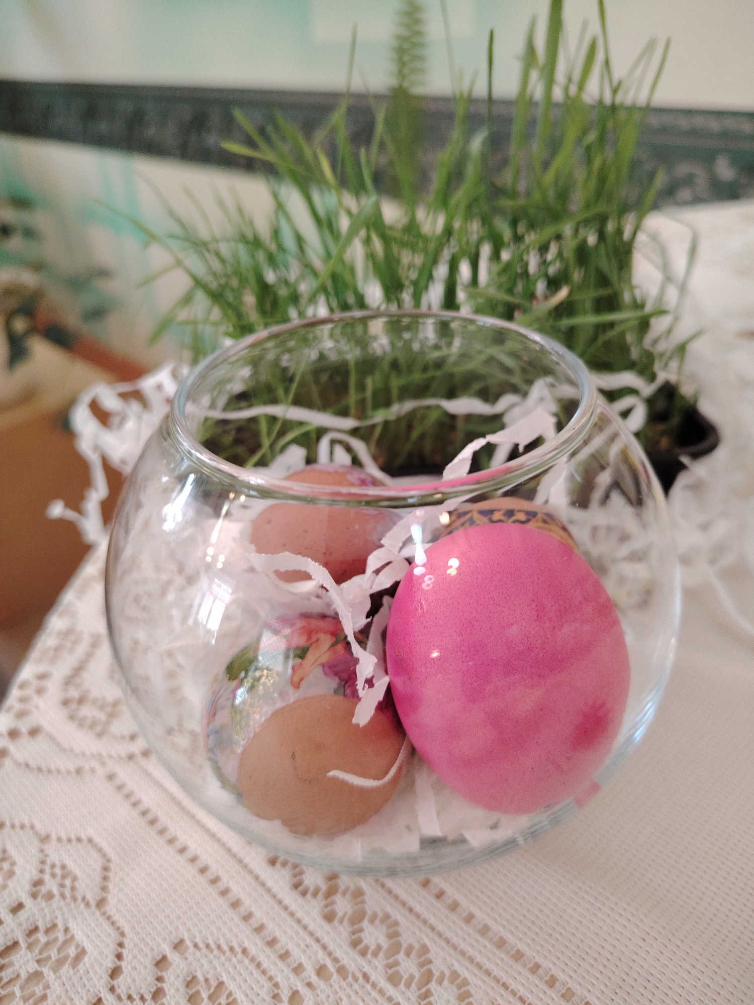 A close-up shot shows a clear glass bowl containing Easter eggs and white paper shreddings, placed on a patterned white tablecloth. Behind the bowl, there's a patch of green grass or wheat sprouts in a pot. The eggs inside the bowl include one painted with floral designs, one brown, and one dyed bright pink. The background is soft and blurred, but hints at a domestic setting. There is no indication of this photo being taken in Sarapul, Russia.