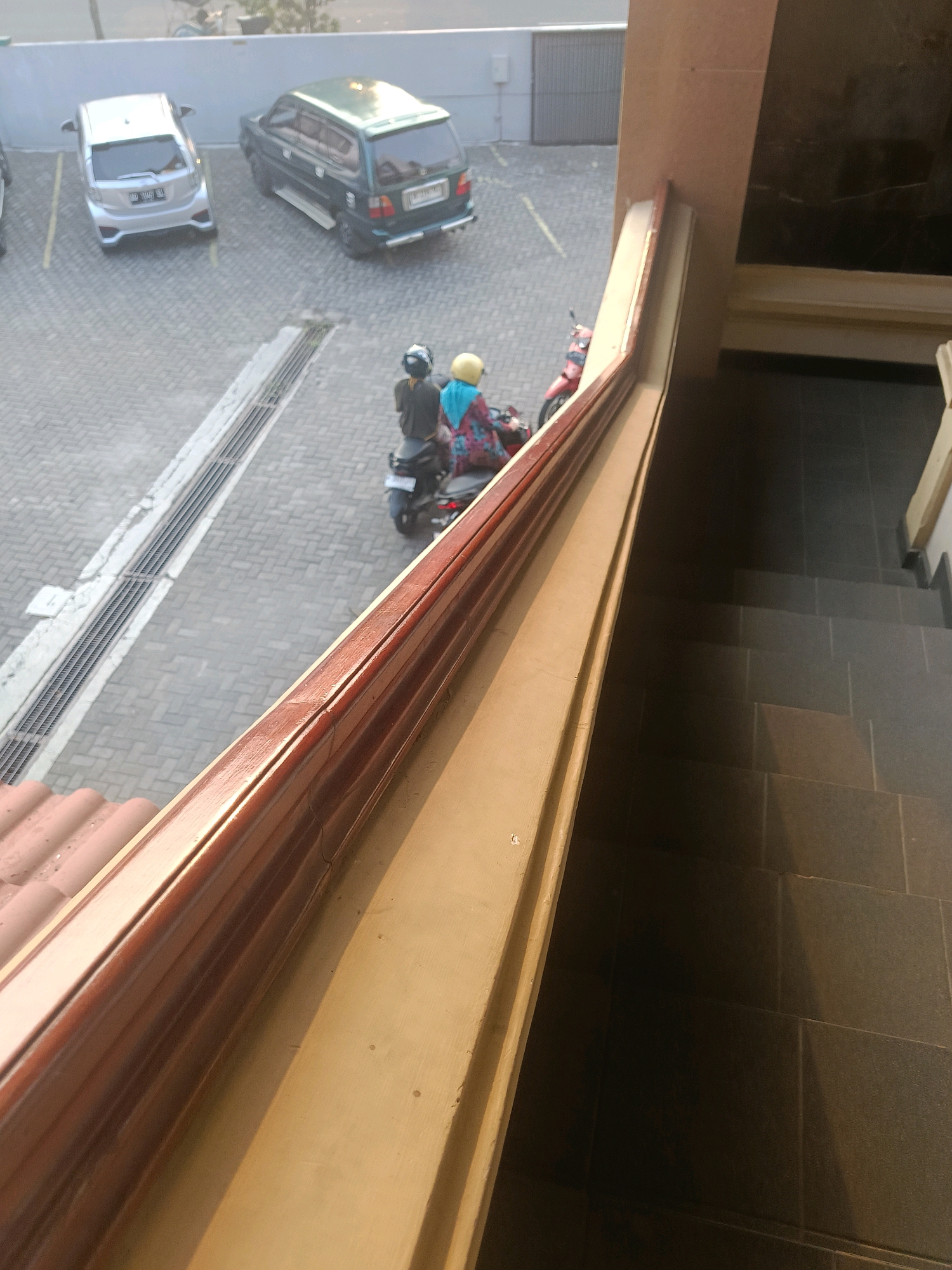 The image depicts an outdoor scene in Yogyakarta, Indonesia, viewed from a slightly elevated perspective looking down. In the midground, two individuals are seated on a dark-colored scooter. The person in the front wears a black helmet and a dark top, while the person behind them has a yellow helmet and is dressed in a vibrant, patterned blue and red garment. The scooter itself is partially obscured.
Further in the midground, two cars are parked on a paved surface composed of interlocking brick pavers. The car on the left is a silver hatchback with its rear facing the viewer. The car on the right is a dark green minivan with its rear also visible. A metal grate runs parallel to the cars and extends into the background.
The foreground and right side of the image are dominated by architectural elements. A brown wooden railing runs diagonally across the frame, casting a shadow. Below the railing, a set of dark gray tiled stairs descends into shadow, with the tiling clearly visible. A tan-colored vertical surface runs alongside the railing. In the upper left quadrant, part of a red-tiled roof is visible. The overall environment suggests a commercial or residential area with parking facilities and outdoor stair access.