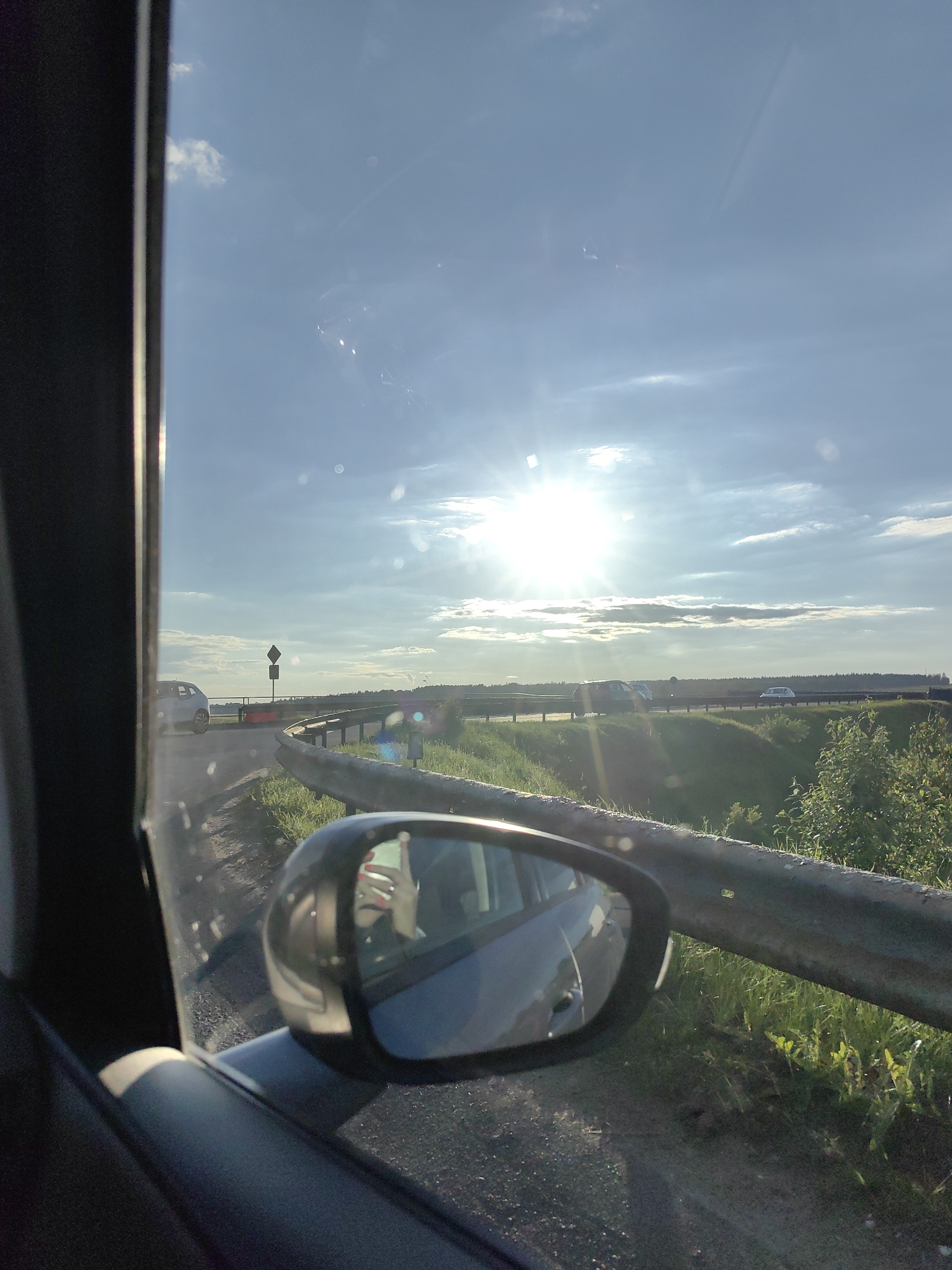 A side-view mirror of a car reflects a hand holding a smartphone, indicating a person is taking a photo or recording a video from inside the vehicle. The car is on a road with a metal guardrail, suggestive of a highway or a rural road. The background shows a bright, sunlit sky with clouds, and a landscape with trees and fields, hinting at an outdoor, possibly rural, setting near the city of Fanipalski Sel'ski Savet, Belarus. Multiple cars are visible on the road in the distance, suggesting it is a moderately busy route. The overall scene depicts a journey on a sunny day.