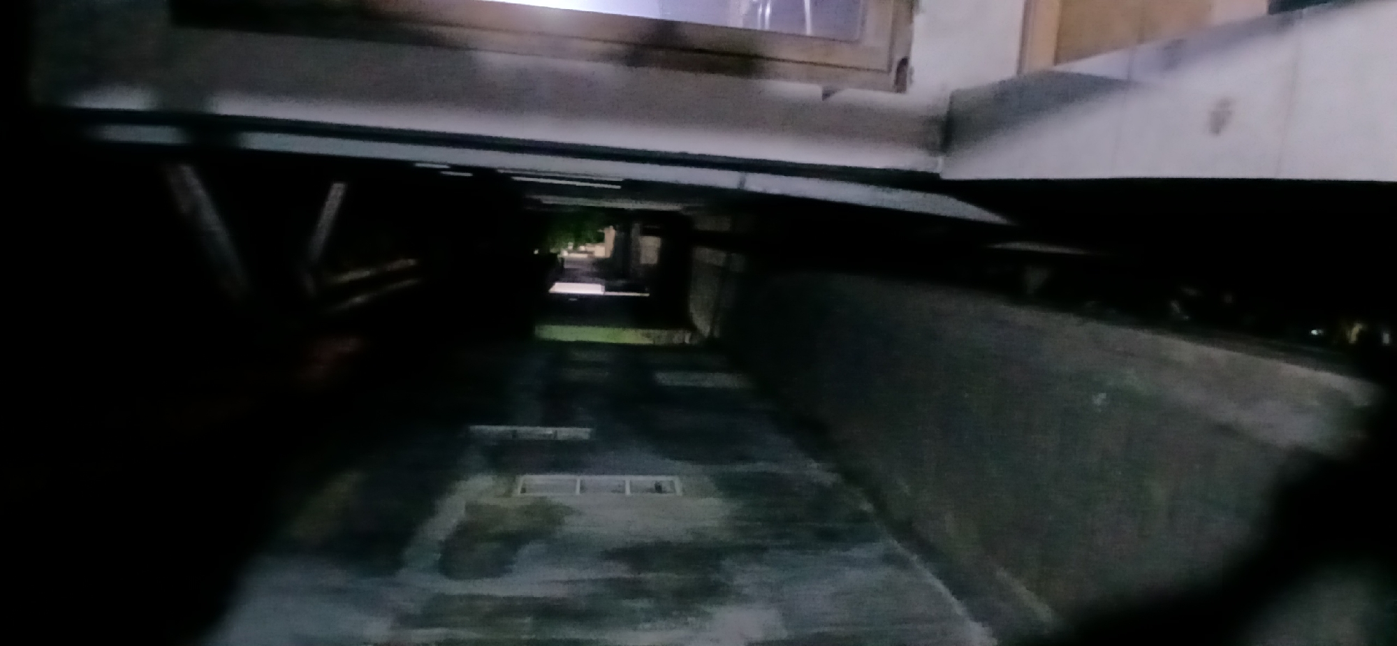 The image shows a dark, narrow alleyway between two buildings at night. The building on the left has a weathered, stained wall with two small, rectangular windows. The building on the right appears to be a balcony with a railing and a window. The alleyway is paved and appears wet, reflecting some dim light. Trees are visible in the distance at the end of the alley. The scene is dimly lit, suggesting it is either a poorly lit area or taken with a low-light camera. The image is grainy and somewhat blurry.