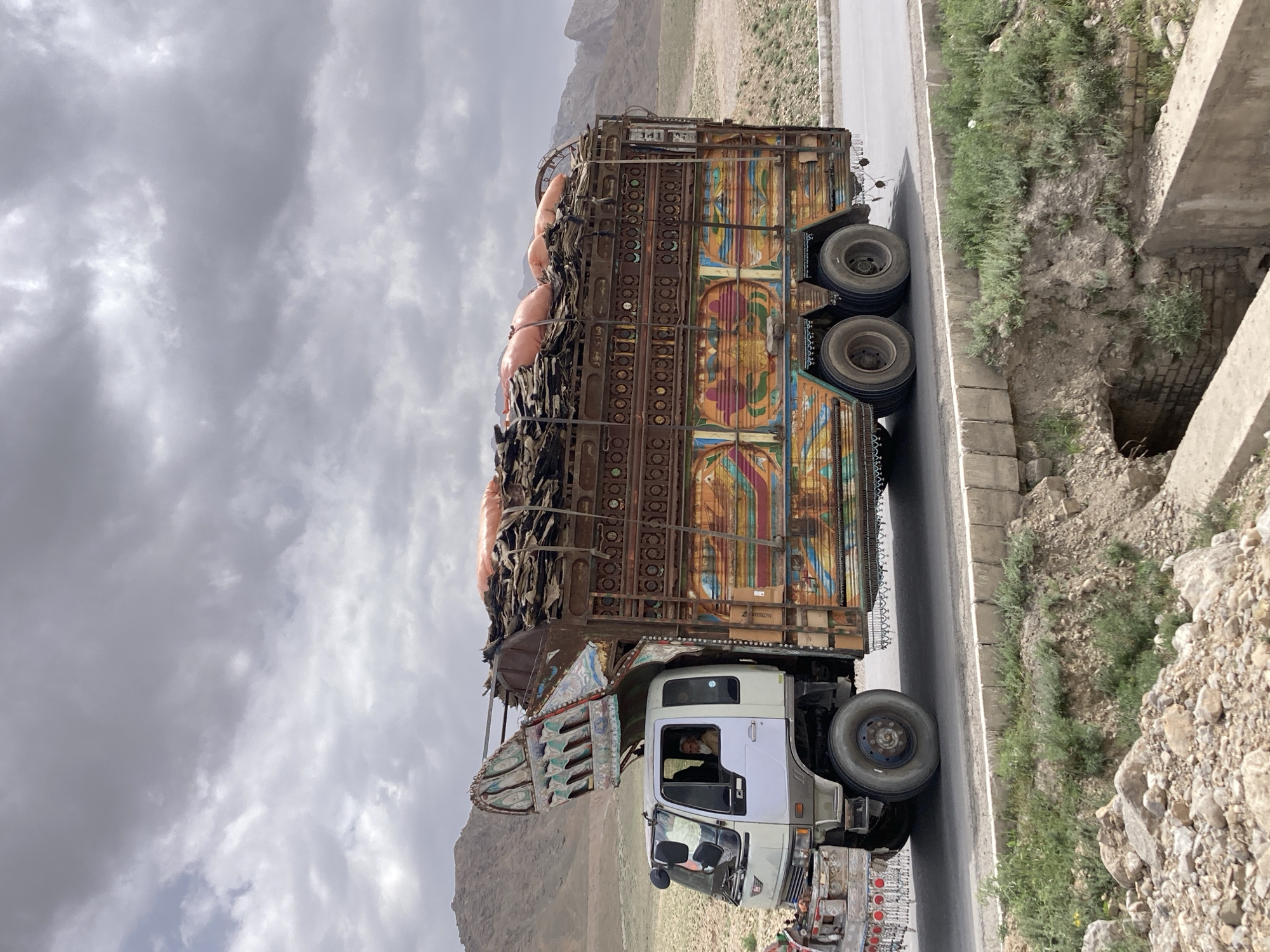 A large, highly decorated truck is captured on a mountain road under an overcast sky in Wulgai, Pakistan. The truck, likely stationary or moving slowly, is laden with a substantial cargo of irregularly shaped, dark bundles and several large, round, orange sacks piled high on its open bed. On the front-left portion of the cargo, the word "محمد" (Muhammad) is visible in white script.
The truck itself is a prominent subject, painted in the vibrant and intricate folk art style common in Pakistan. Its sides feature colorful geometric patterns, floral motifs, and mandalas in hues of orange, blue, green, and red, accented by elaborate metalwork with cut-out designs. The cab of the truck is white and light gray, and a person, presumably the driver, is visible through the open window, looking towards the camera.
The setting is a rugged, mountainous landscape with sparse, dry vegetation and rocky terrain. The truck is positioned on a paved road, next to which is a dirt shoulder with some green plants, leading to a visible ditch or culvert with concrete and brick structures. The sky is largely covered with thick, gray clouds, suggesting an overcast day. No direct sunlight is apparent. The overall impression is one of a working vehicle in a remote, challenging environment. Faint inscriptions in Urdu or a similar script are present on various parts of the truck's decorated body, adding to its unique character.