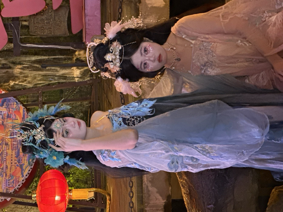 The image features two young females in elaborate traditional Chinese-style attire, posed outdoors at night in Yuzhong District, China. The individual on the left stands, facing slightly right, with her right hand touching a forehead jewel. She wears a blue and black garment with shimmering embroidery, a transparent light blue overlay, and an intricate blue feathered and silver headdress. Glitter makeup accents her eyes and forehead. To her immediate left, a large, illuminated red traditional Chinese lantern with a yellow stand is visible. The individual on the right is partially visible, appearing to be seated or kneeling, facing forward. She wears a pink embroidered dress, a silver and pearl headdress with pink floral accents, and a silver necklace. Her face also features prominent pink glitter makeup around the eyes. The background includes a decorative screen or wall with traditional Chinese architecture designs, an illuminated vertical water feature resembling a waterfall, and a dark bell-shaped object. Dark metal chains traverse the midground horizontally. The scene is illuminated by artificial lighting, creating a distinct visual atmosphere.