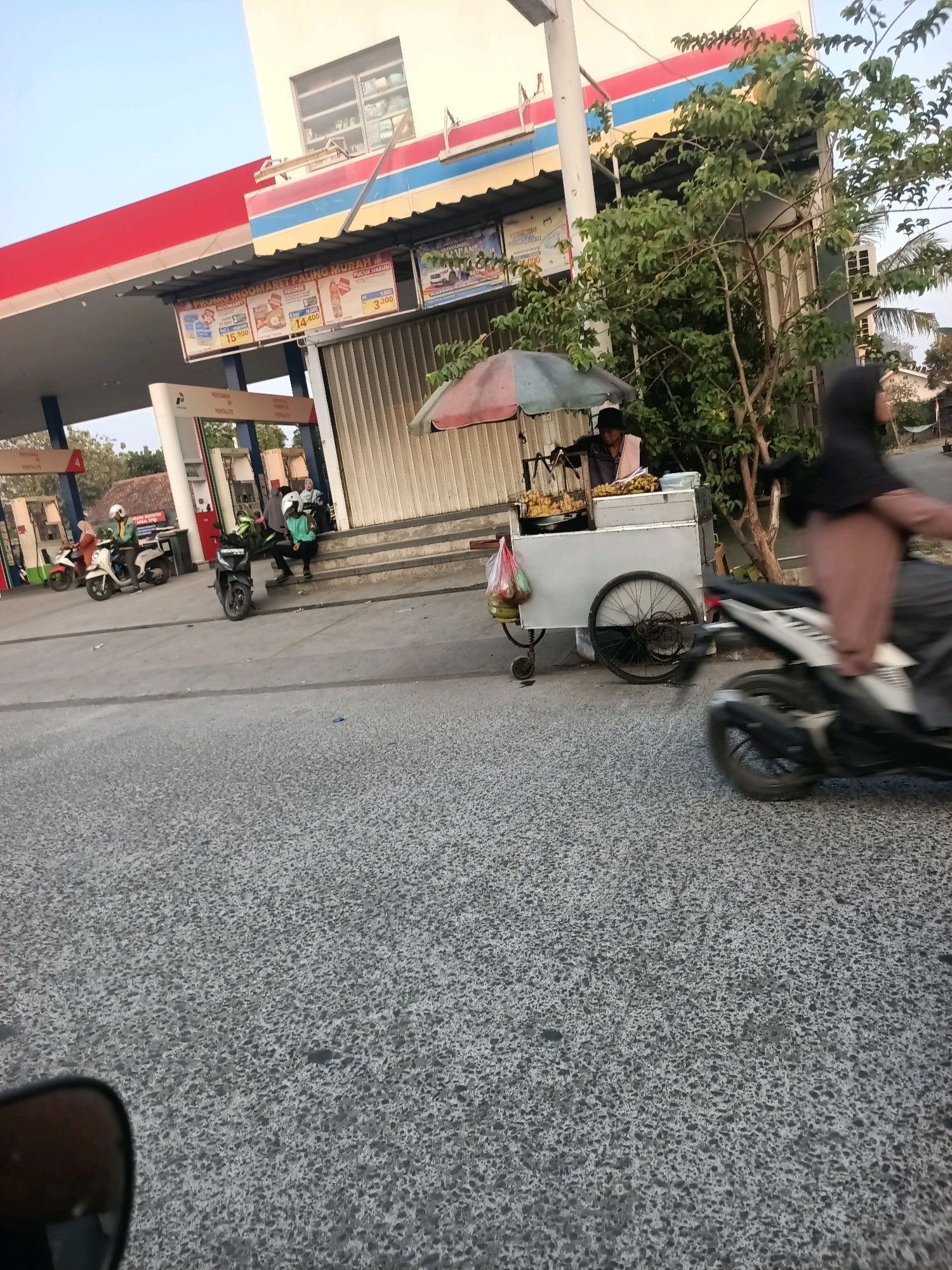 The image depicts a street scene in Wanajaya, Indonesia. A gas station with prominent red and blue signage is visible, along with a convenience store. In the foreground, a person on a motorcycle is captured in motion, their form blurred by the speed. To the left, several other motorcycles are parked, and people are standing or sitting near the gas station pumps. A food cart with a striped umbrella is situated near the entrance of the convenience store, with a vendor attending to it. The street is paved with asphalt, and there is a grassy area and trees to the right of the food cart. The sky is clear and blue.