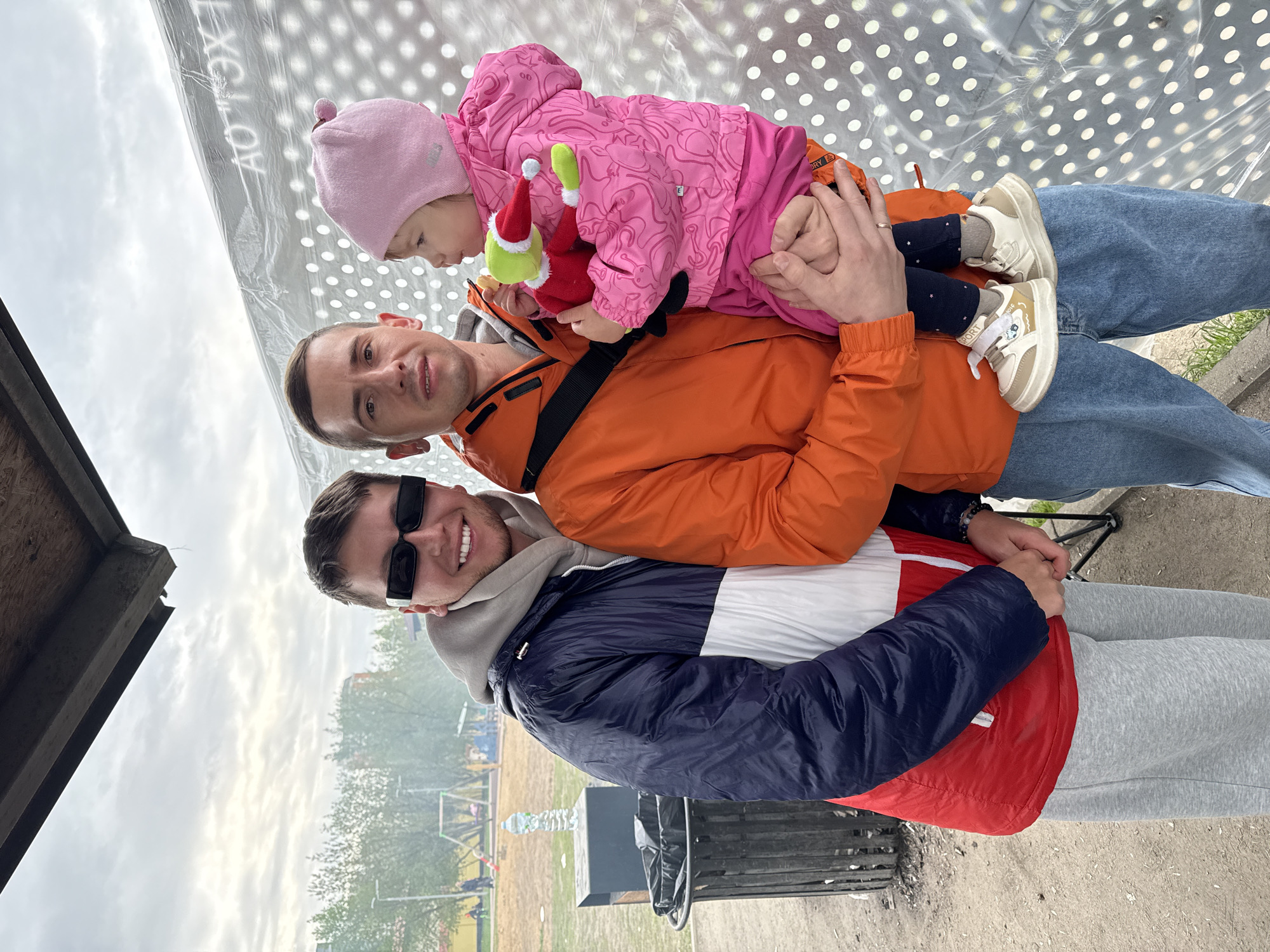 The image shows three individuals. A man wearing sunglasses sits in a folding chair; atop him sits another man, who in turn supports a child. The child wears a pink snowsuit and clutches a stuffed toy. The man in sunglasses wears a navy, white and red jacket and gray sweatpants. The middle man is dressed in an orange jacket and blue jeans. They are outside in a park. In the background are trees and a playground. The sky is overcast. The location could be Moscow, Russia.