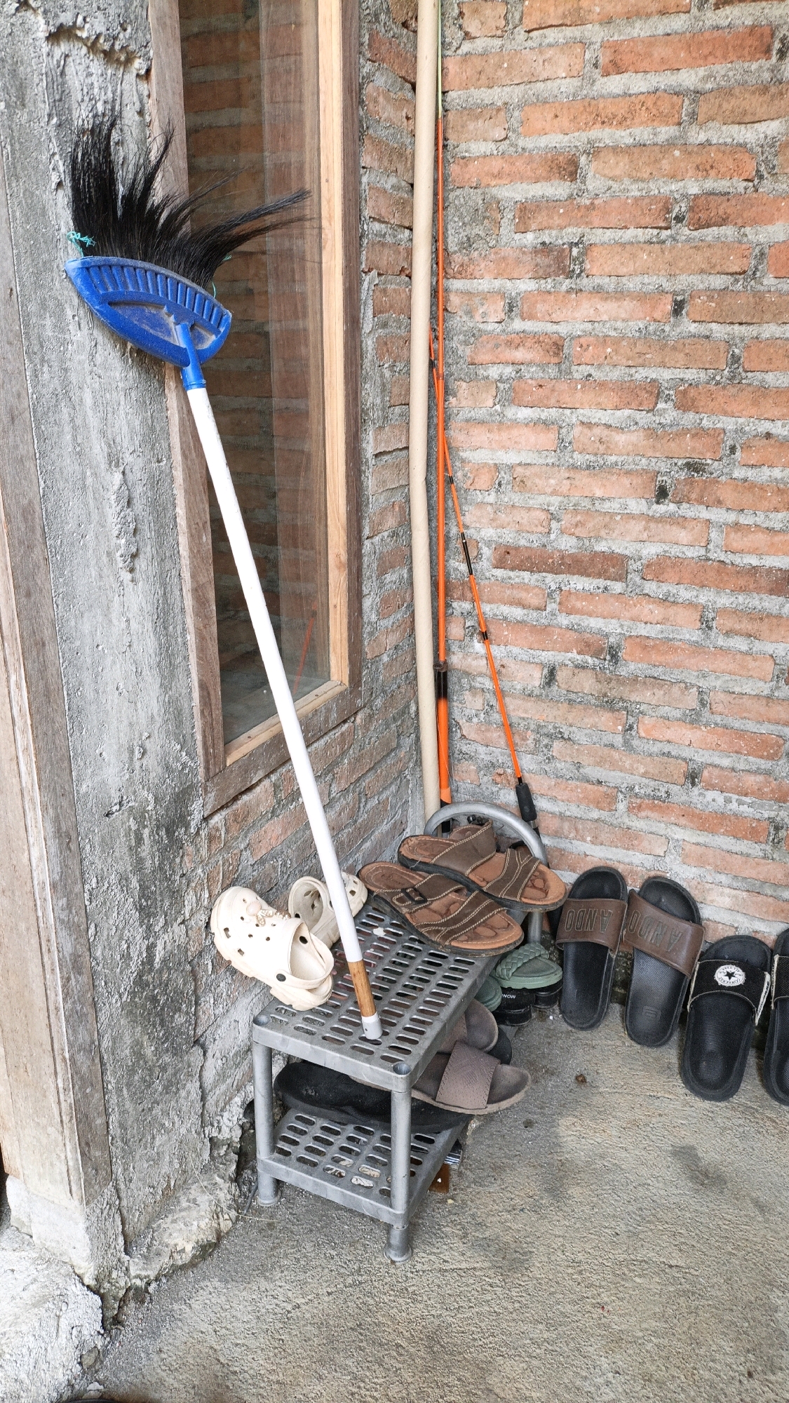 In Cikal, Indonesia, outside a building with a textured, light gray concrete wall, a wooden window frame with a glass pane, and a red brick wall, a collection of cleaning tools and footwear is arranged. Leaning against the wall is a blue-headed broom with a white handle, positioned to the left of the frame. To the right of the broom, two long poles are propped up: one is a light beige color and the other is bright orange. In front of the poles, a small, gray, two-tiered plastic shelf holds several pairs of sandals and clogs. On the top shelf, a pair of light beige clogs are visible on the left, followed by a pair of brown strap sandals and a pair of dark green flip-flops. On the bottom shelf, a pair of dark brown strap sandals rests on the right side. Further to the right, on the concrete floor, a line of black slide sandals is neatly arranged. The overall scene suggests an outdoor storage area for household items.