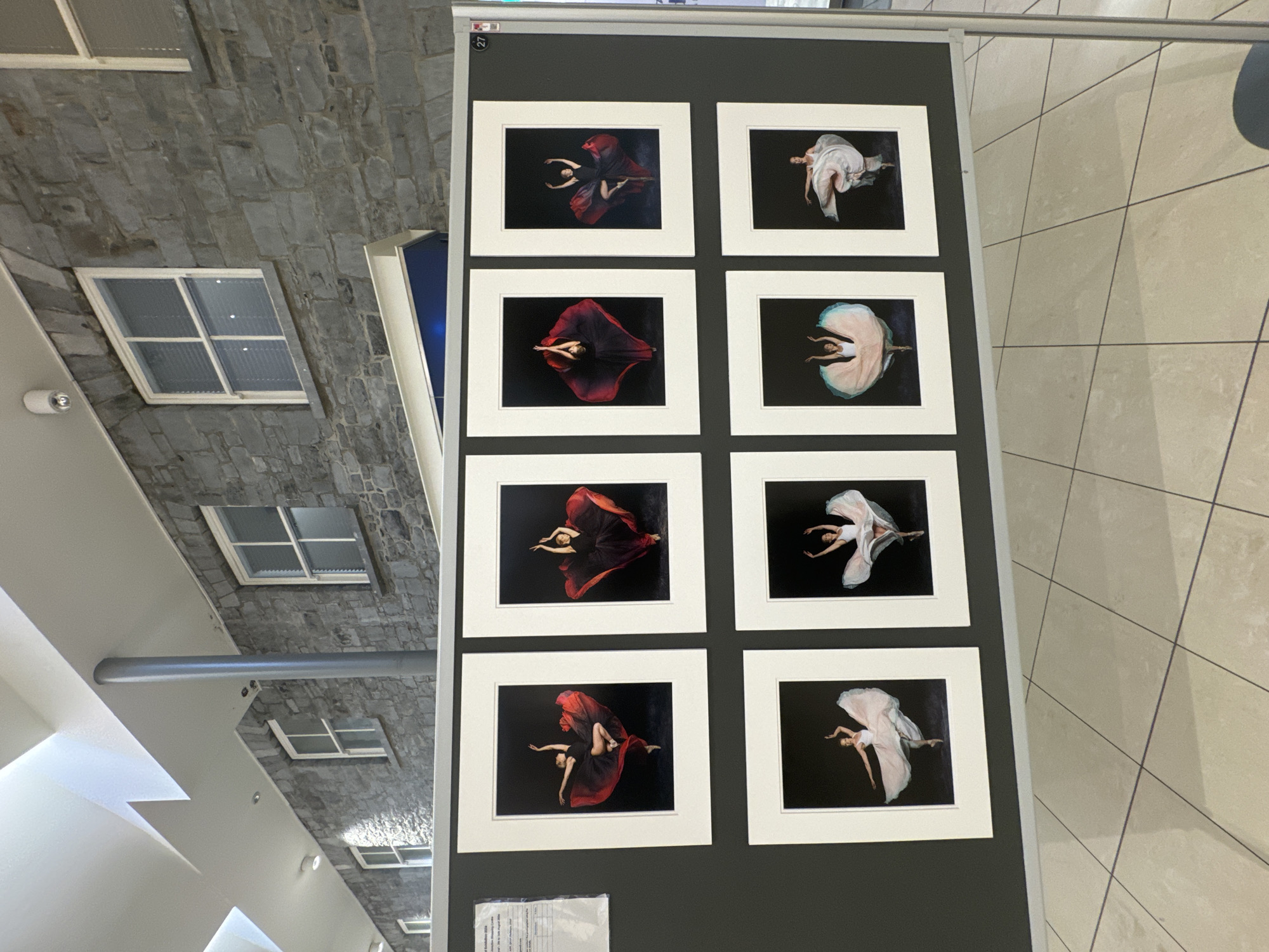 An exhibition display features eight framed photographs of ballerinas against black backgrounds. The photographs are arranged in two rows of four. The top row shows ballerinas in red or dark red skirts, and the bottom row shows ballerinas in lighter colored tutus or skirts. The display is set up in a modern building with stone walls and large windows. The floor is tiled. The image appears to be taken indoors, possibly in a public space like a gallery or a community center, as suggested by the display structure and the architecture of the building. The presence of a numbered label '27' on the right side of the display suggests it is part of a larger exhibition. The lighting appears to be artificial. The scene is static and depicts an art exhibition. Given the subject matter of the photographs and the architectural style, this could be a display at the Kilkenny Arts Festival or a similar cultural event in Kilkenny, Ireland, known for its historic architecture.
