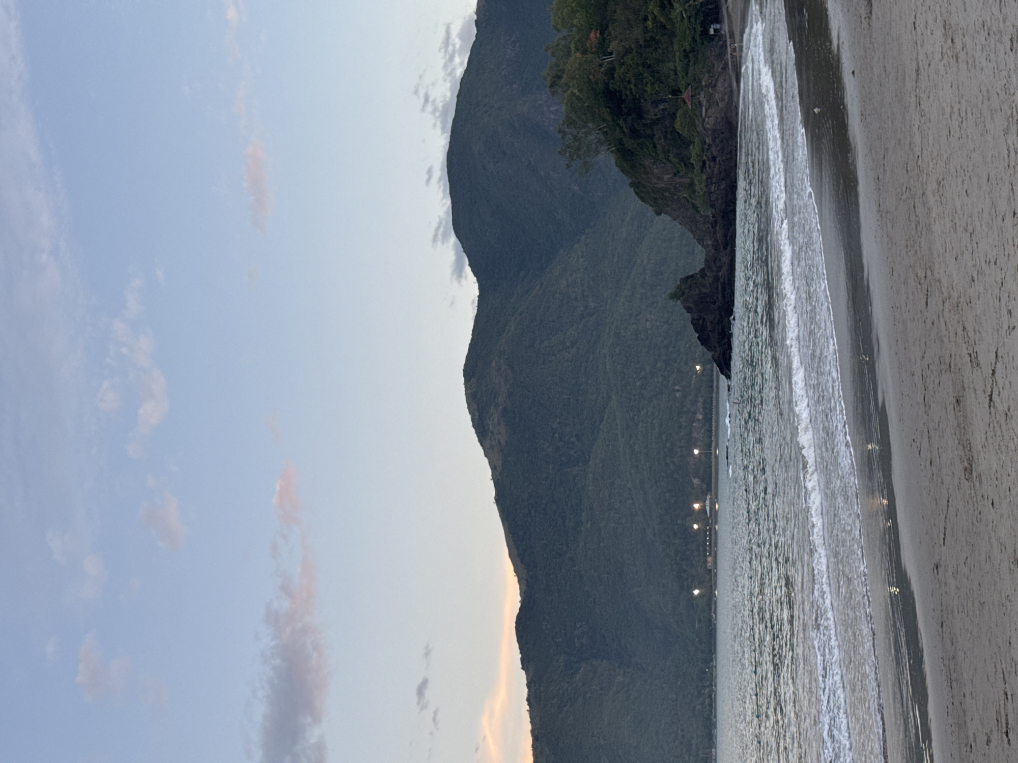 The image depicts a coastal scene at dusk or dawn in Nha Trang, Vietnam. A sandy beach occupies the foreground, with gentle waves lapping at the shore. The ocean stretches to the horizon, meeting a range of lush, green, tree-covered mountains. The sky is a soft gradient of pale blue and pink hues, with scattered clouds. In the middle ground, a rocky outcrop juts into the water, topped with dense vegetation. In the distance, a line of lights is visible along the coastline, suggesting a populated area or resort. A few faint figures can be seen on the beach to the right of the frame. The overall impression is one of tranquility and natural beauty.