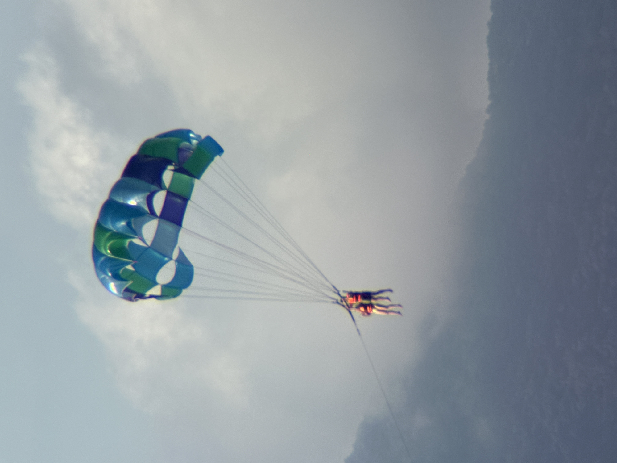 A parasail with blue and green panels flies in the sky above a blurred, mountainous terrain that could be Đà Nẵng, Vietnam. Two people wearing orange life vests are suspended beneath the parasail, holding onto the harness. They appear to be enjoying a recreational activity. The sky is overcast with soft clouds.