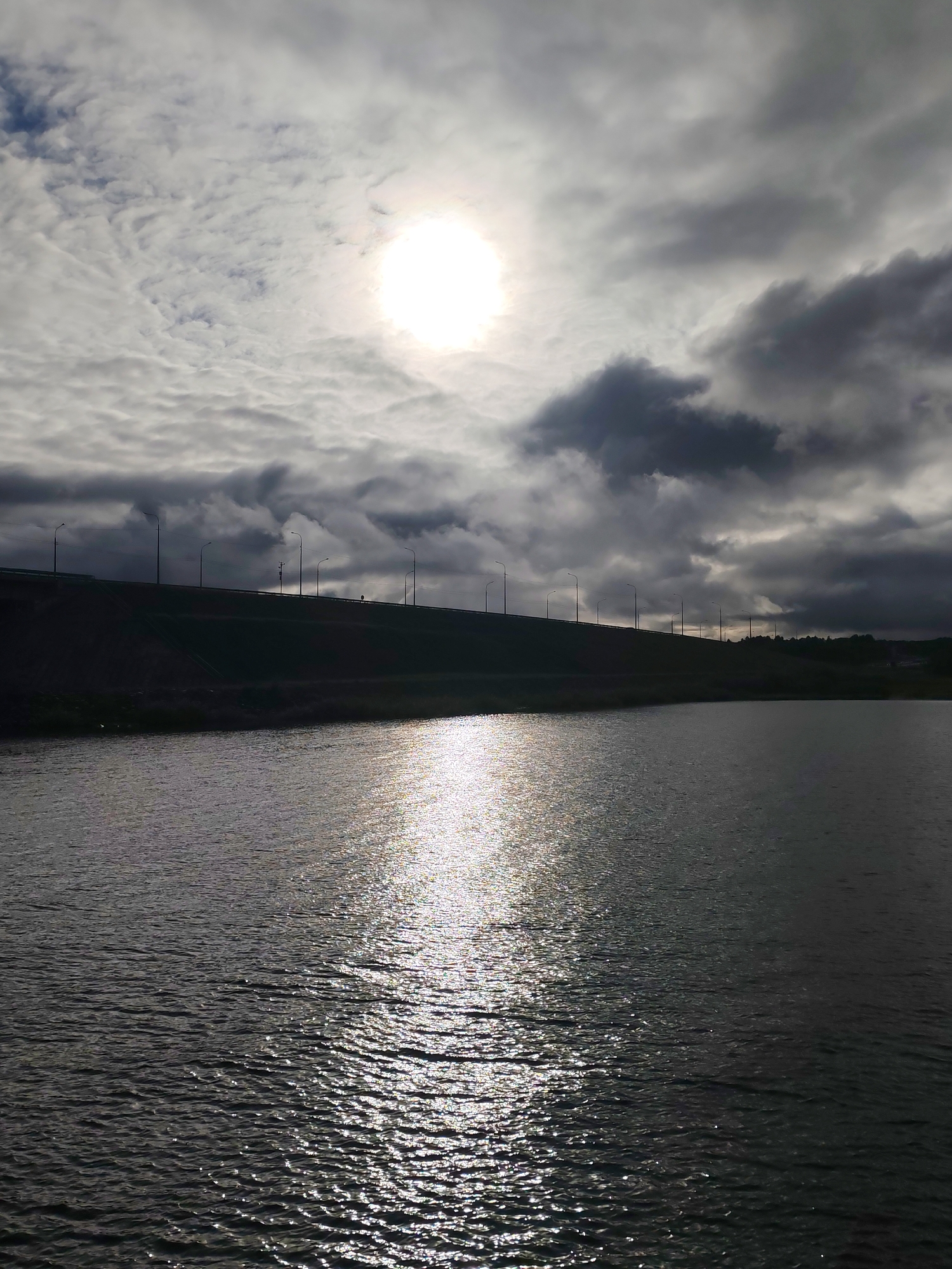 A medium-shot captures a body of water under a sky filled with mixed white and dark gray clouds, where the sun shines brightly. A bridge or overpass can be seen in the distance, supported by evenly-spaced lamp posts along its length. The lighting conditions suggest a cloudy day with the sun trying to break through. The water surface reflects the sunlight, creating a shimmering effect. The location is Нифантово, Russia.