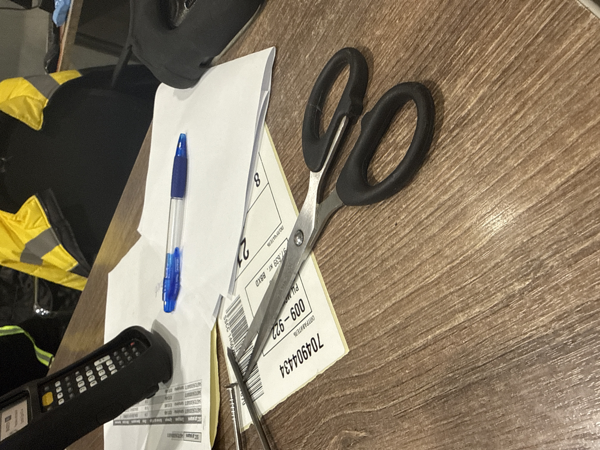 A pair of silver and black scissors, a blue ballpoint pen, and a black barcode scanner rest atop a stack of white papers with printed Russian text and numbers on a light brown wooden surface. The text on one of the papers includes "704904434", "ОПТРАВИТЕЛЬ", "009-922", "97.639 кг. BBXD", "ПОЛУЧАТЕЛЬ", "21", and "8". Part of a yellow and black high-visibility garment is visible to the left, along with dark-colored items that are out of focus. There's no information given or visible that allows for determination of the location in the city of Lupanovo, Russia.