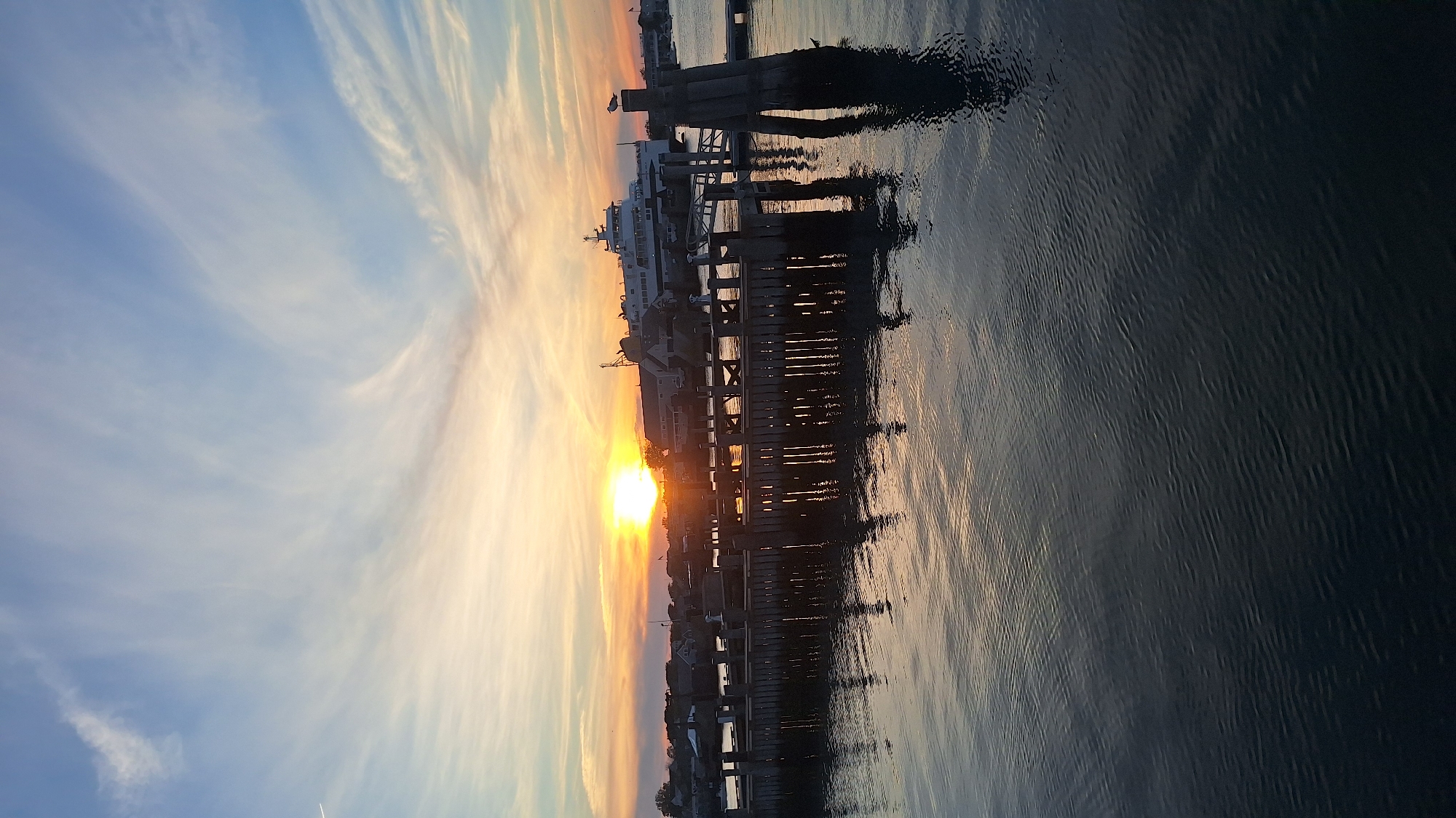 This is a photograph taken at sunset on Nantucket, Massachusetts, USA, featuring a pier and a body of water. The sky displays a gradient of colors from pale yellow near the horizon to a soft blue higher up, with wispy white clouds. The sun, a bright orb, is setting behind a cluster of buildings and a large ferry docked in the distance. The foreground is dominated by the calm, dark water, reflecting the light from the sky and the structures on the pier. The pier itself is made of dark wood and is supported by sturdy posts, with a few more posts visible in the water. A seagull is perched on one of the posts on the right side of the image. The overall scene evokes a peaceful, maritime atmosphere typical of coastal towns.