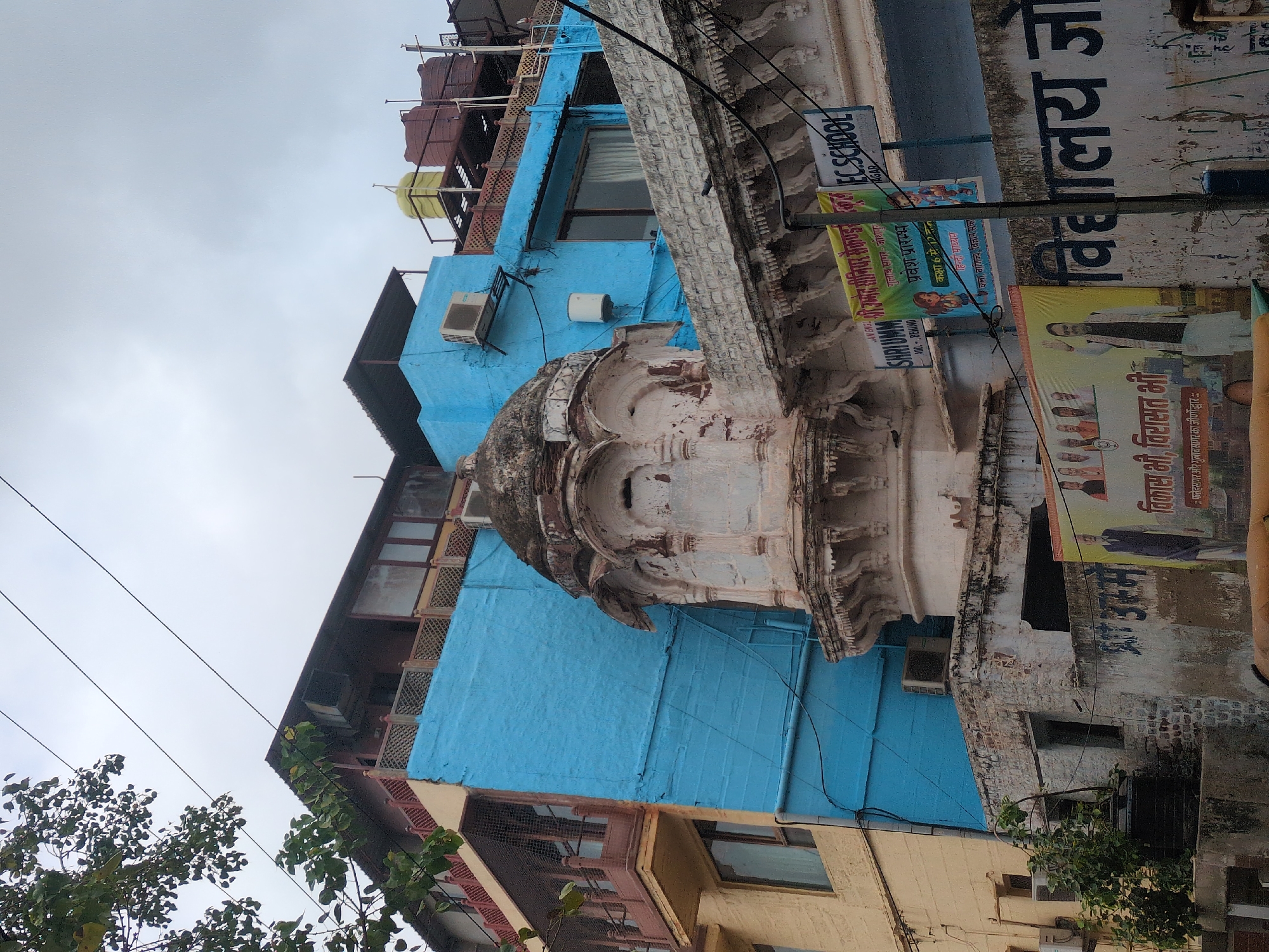 This is an exterior, eye-level, medium shot captured on an overcast day in Jodhpur, India, focusing on the exterior of buildings with a mix of architectural styles. The foreground features a textured, weathered white wall with decorative carvings along its top edge. Below this, a large banner displays images of people and text in Hindi, alongside another banner advertising "EC SCHOOL". A vertical pole partially obscures the banners and extends upwards.
Dominating the mid-ground is a light blue painted wall, appearing to be a modern addition or renovation, with an air conditioning unit mounted on it. Adjacent to this is a section of an older building painted in a pale yellow, featuring wooden balconies with intricate latticework and metal grates. A small, ancient-looking domed structure, made of weathered stone with intricate carvings, sits prominently in front of the blue wall, appearing to be part of an older, possibly religious or historical, building. Green foliage from a tree hangs from the upper left corner, with power lines crisscrossing the sky. In the background, on a rooftop, are several large water tanks, one yellow and two dark reddish-brown, suggesting the location is likely in a densely populated urban area where water storage is common. The overall impression is of a neighborhood where different eras of architecture coexist.