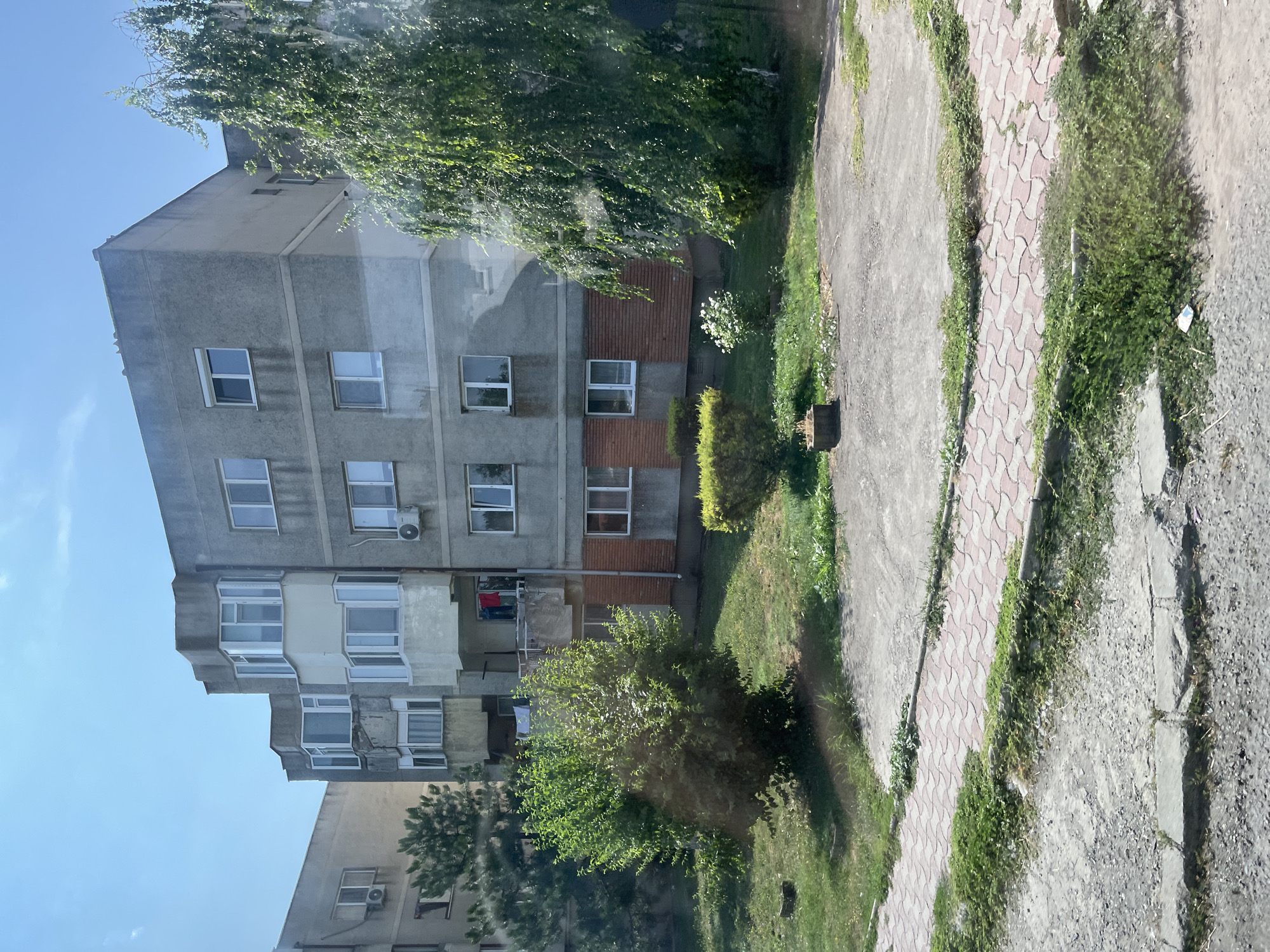 The image depicts an apartment building in the city of Negru Vodă, Romania. The building is made of concrete and features multiple stories with numerous windows. Some of the windows are bay windows, and there are balconies visible, some with laundry hanging. The building's facade is gray and shows some signs of wear. In front of the building, there is a grassy area with some trees and bushes. A paved walkway with red and gray interlocking pavers runs through the scene, bordered by grass. The overall environment appears to be a residential urban setting.
