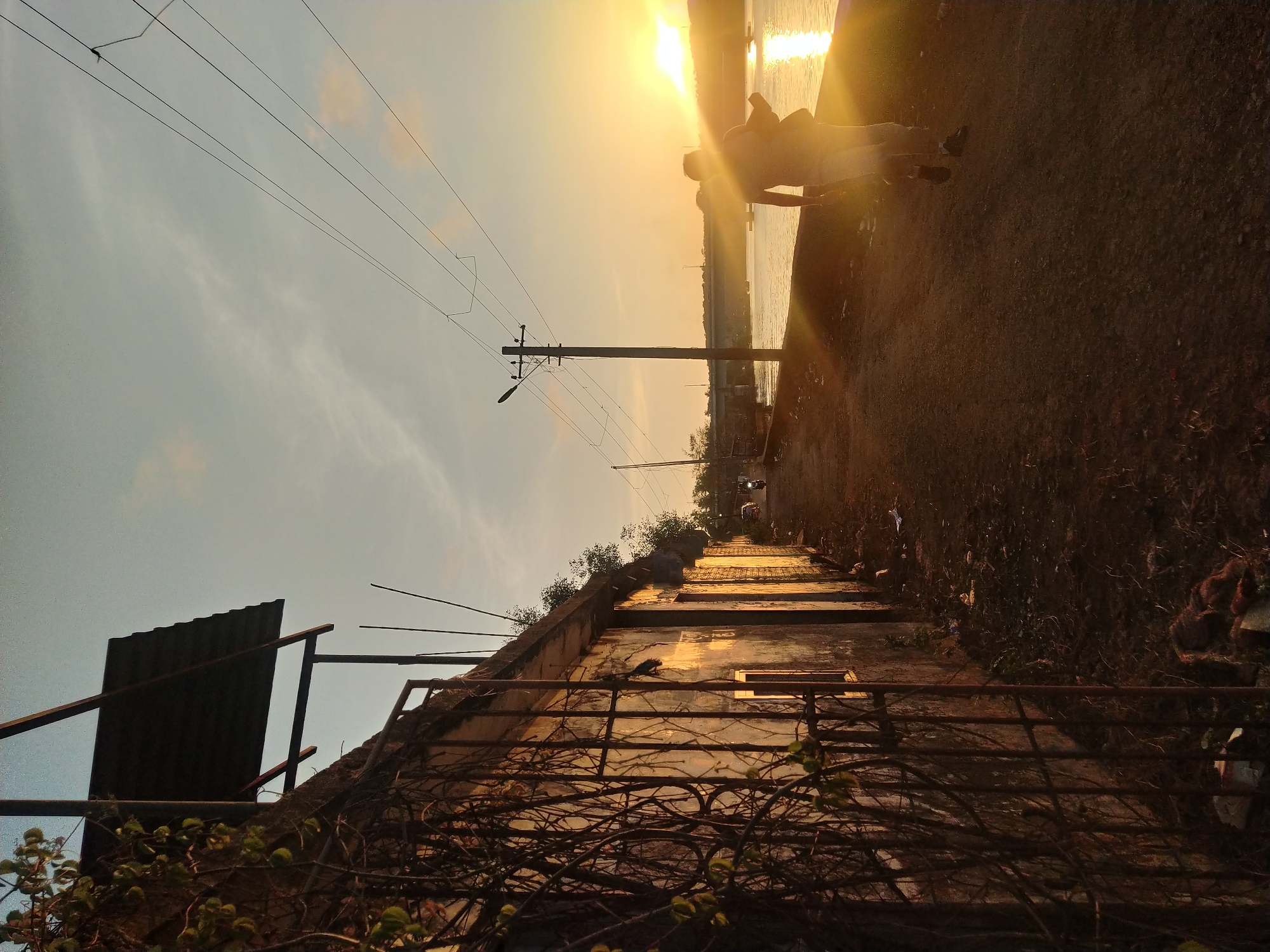 The image shows a sunlit scene in Oxel, India. In the background, two people are silhouetted against the bright sun reflected on a body of water, possibly a river or the sea. An electrical pole stands to the left of the people, with power lines stretching across the sky. The foreground features a weathered structure, perhaps a building or a retaining wall, with a rusted metal fence covered in vines. The structure is bathed in sunlight, creating a warm, golden hue. The overall perspective suggests the photo was taken from a slightly elevated position, looking down towards the water.