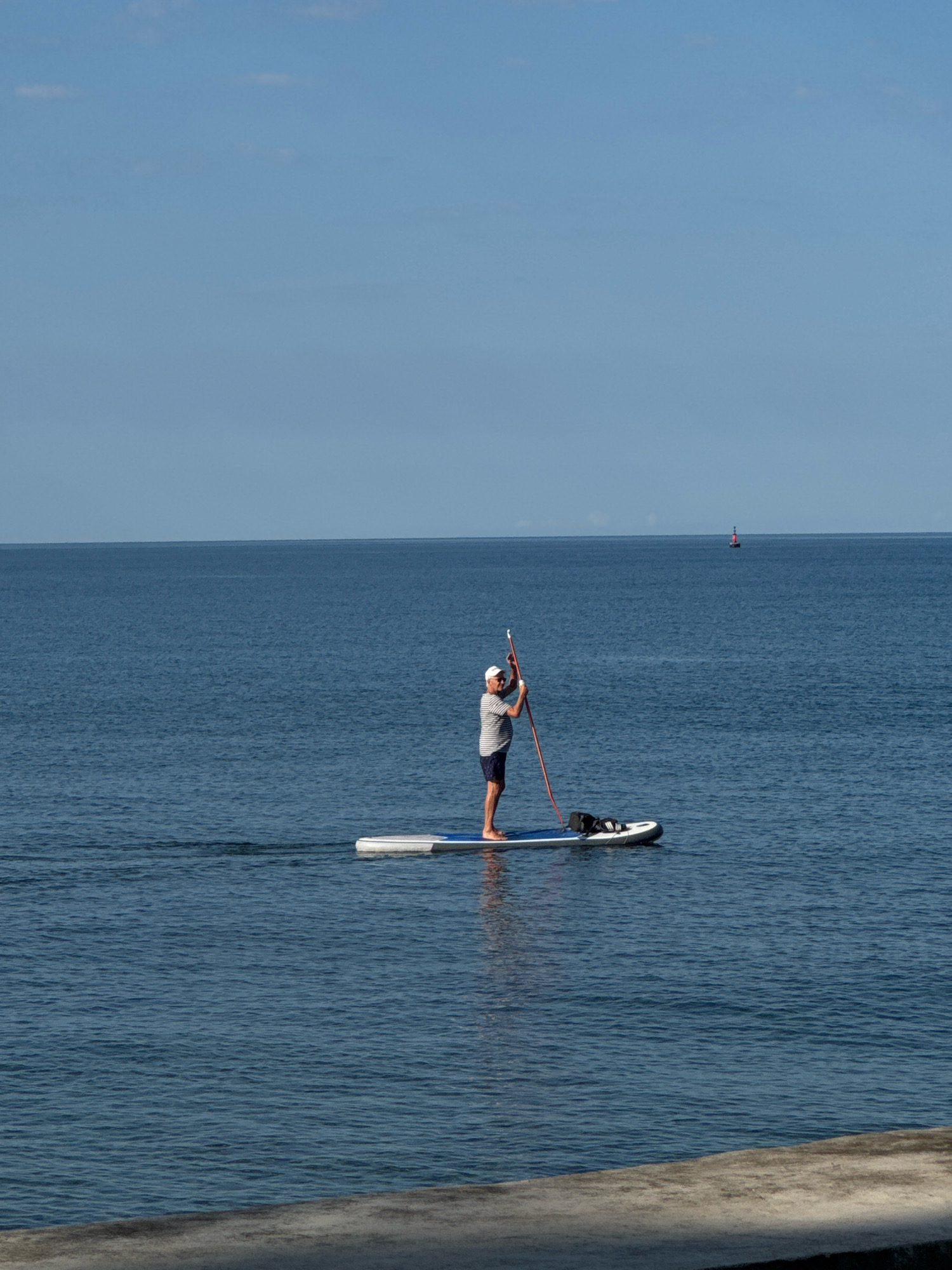 A man is paddleboarding on a calm, deep blue body of water under a clear sky. He is wearing a white cap, a horizontally striped t-shirt, and dark shorts. He is holding a paddle and appears to be moving forward. A black bag is secured on the paddleboard in front of him. In the distance, a red and black buoy is visible on the horizon. The foreground shows a concrete embankment, suggesting the presence of a shoreline or harbor area. The scene is likely set in or near the city of Novigrad, Croatia, given the calm waters and the presence of navigational markers typical of coastal towns.