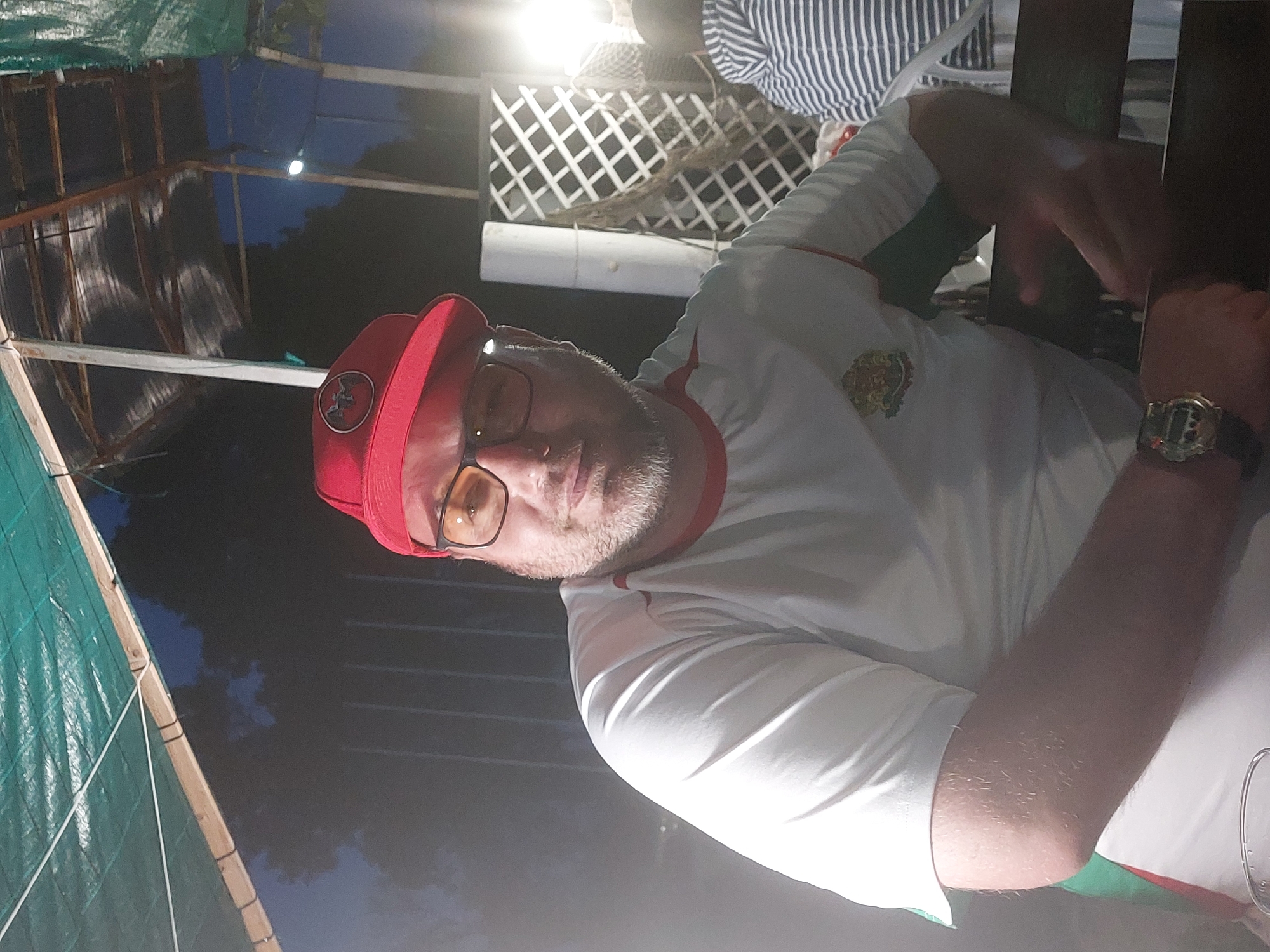 The photograph appears to be taken outdoors, likely at night, in Tsarevo, Bulgaria. A man is the primary subject. He wears a red baseball cap with a dark emblem, glasses, and a white t-shirt with a red trim around the neck. A wristwatch is visible on his left wrist. He is positioned at a table, potentially in a restaurant or bar setting. The background includes a white trellis structure and a striped shirt is partly visible. A partially-covered structure, perhaps a tent or awning, is visible in the periphery. The overall scene seems casual.