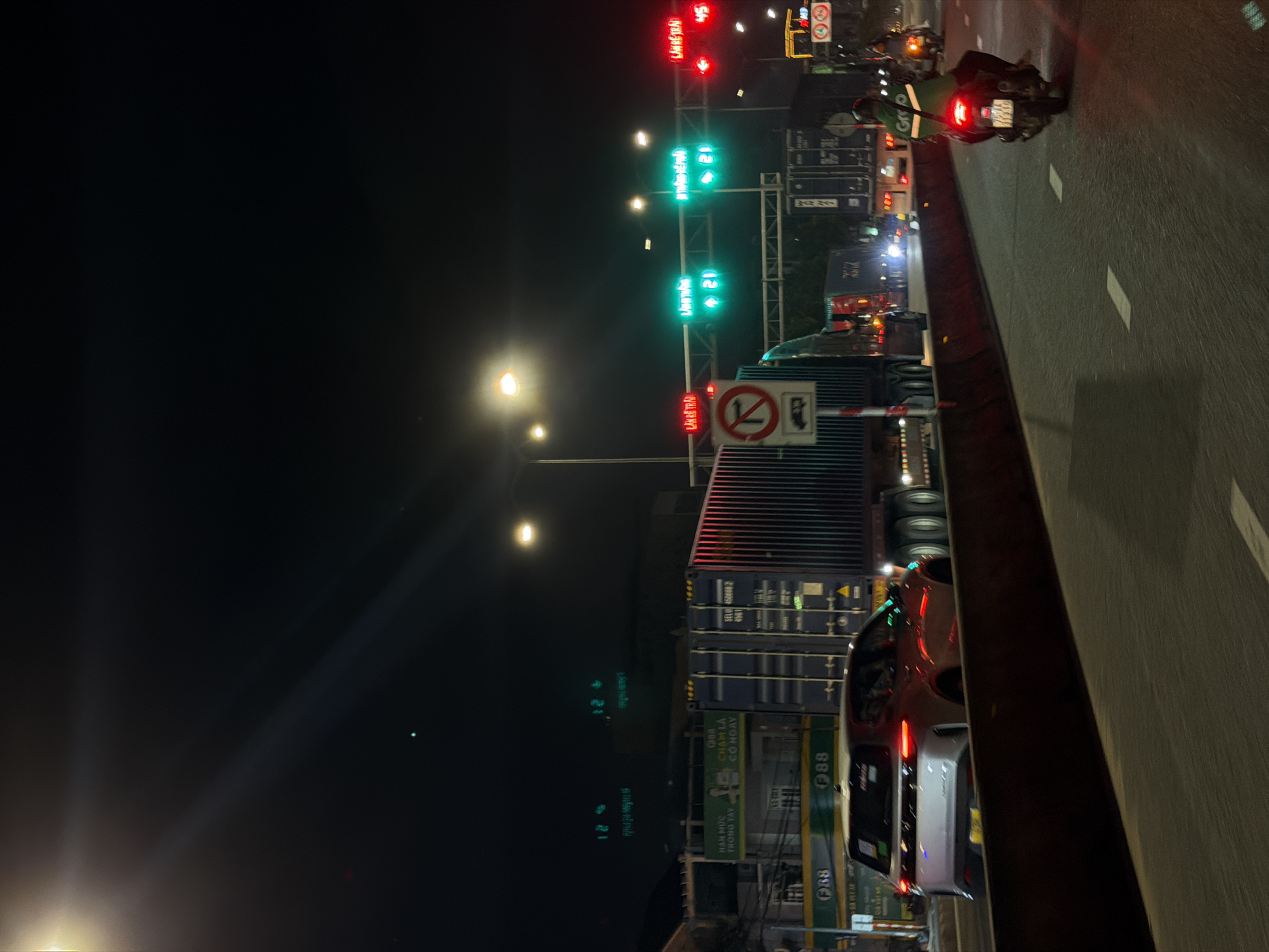 This is a night-time photograph taken from a vehicle looking forward and slightly to the right, capturing a highway or major road scene in Đà Nẵng, Vietnam. The focus is on traffic lights, road signs, and various vehicles.
In the foreground, a red car is visible, its headlights illuminated. To the left, a truck with several containers is stopped. Further back, a motorcycle rider wearing a bright green vest is seen. Traffic lights with red and green signals are prominent, some displaying numbers like "21" and "12," possibly indicating lane guidance or speed limits.
Several road signs are visible, including a circular sign with a red border and a diagonal line through a symbol, indicating a prohibition. Other signs contain Vietnamese text, such as "HẠN MỨC TRONG TAY" and "CHẠM LÀ CÓ NGAY." The overall atmosphere is that of a busy urban nocturnal environment, with the artificial lights of the city and vehicles illuminating the road. The time of day is clearly night, and the presence of traffic suggests ongoing activity despite the darkness. The road surface is wet, hinting at recent rain or ongoing light precipitation.