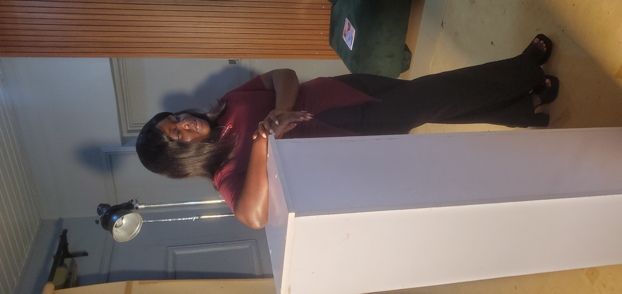 A Black woman with dark hair and a maroon top is leaning against a white pedestal in a room. She is wearing dark pants and black platform heels. Her left arm is resting on the pedestal, and her hands are clasped in front of her. The background includes a metallic lamp on a stand, a wooden wall panel, and a dark green bench with a magazine on it. The floor is a light yellow color. The woman appears to be looking down and to her right. The room is lit by artificial light.