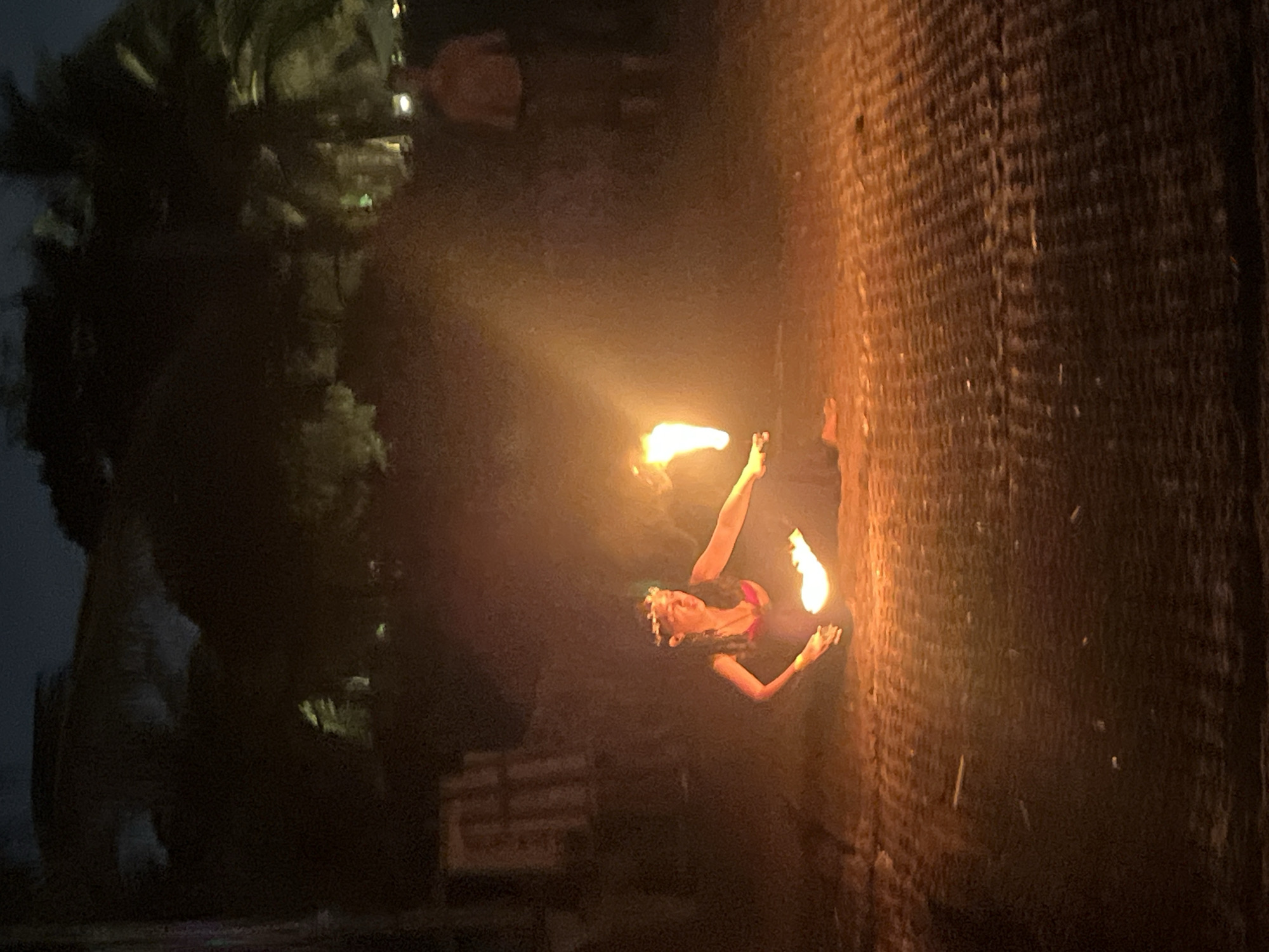 A young woman is performing fire dancing on a woven mat at night. She is wearing a flower crown and a pink bikini top with a dark skirt. Flames are visible from her hands and a torch. In the background, palm trees and a few blurry figures are visible. This scene appears to be a cultural performance, possibly at a resort or beachside venue. The location is not identifiable within the city of Đà Nẵng, Vietnam from the image alone.
