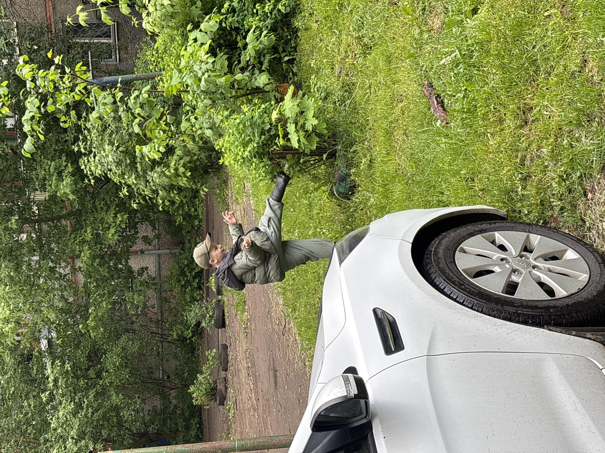 The image, taken in Заволжье, Russia, depicts a white car parked on the edge of a green grassy area. A person wearing a green jacket, hat, and dark footwear is standing near the front of the car. The background includes trees, bushes, and a building with visible windows. The ground appears to be a mix of grass, dirt, and some paved areas. There is also a section of tires lining the edge of a path or road.
