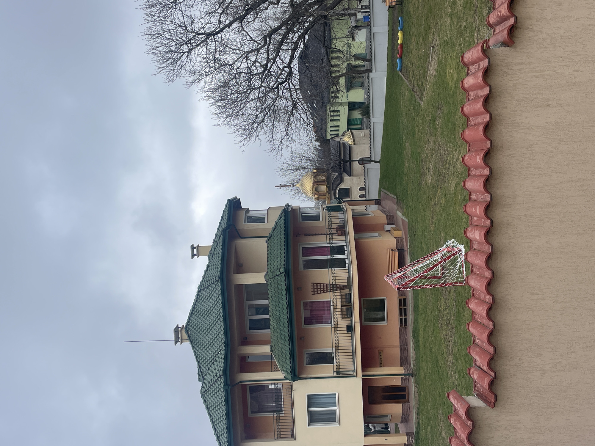 The image shows a multi-story building with a green tiled roof in Sofia, Bulgaria. Adjacent to the building is a lawn area featuring a soccer net and a church with a golden dome. The weather appears overcast. A tree with bare branches is also visible, and a building is behind it, adjacent to a playground with yellow and blue rings. The photo appears to have been taken from an elevated position on the roof of the right adjacent building.