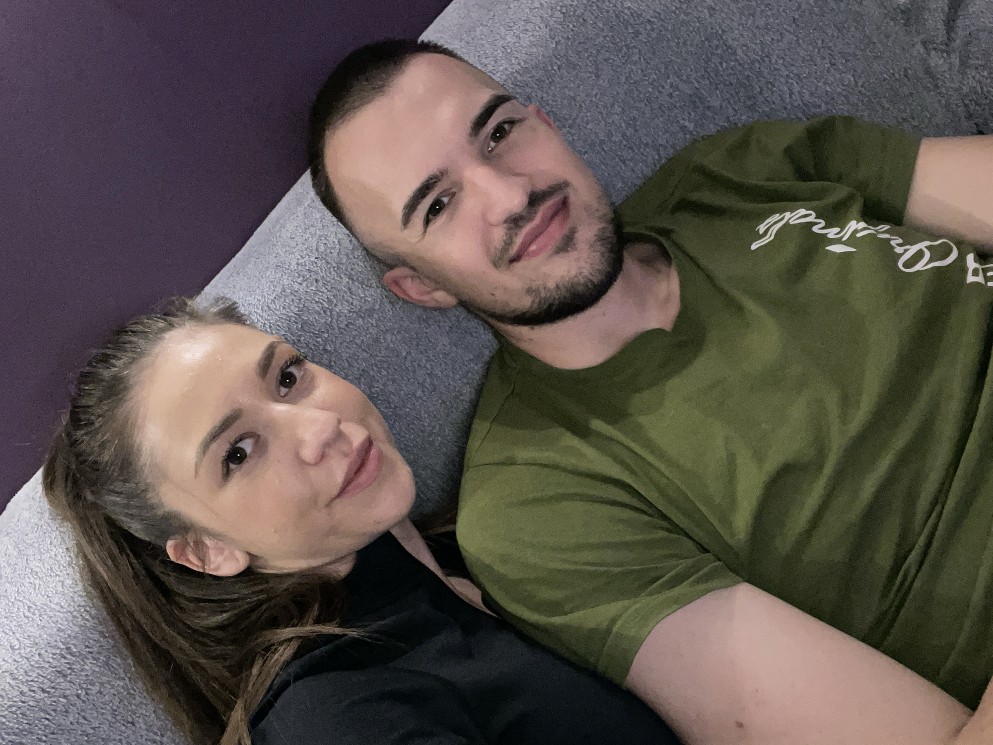 Two individuals are lying on a grey sofa against a purple wall. The man is closer to the right edge of the image, wearing a green t-shirt with white writing on it. He has short dark hair and a beard. The woman is on the left, wearing a black top, with her brown hair pulled back in a ponytail. The image is shot from above. There is no specific indication that this scene is in Sofia, Bulgaria.