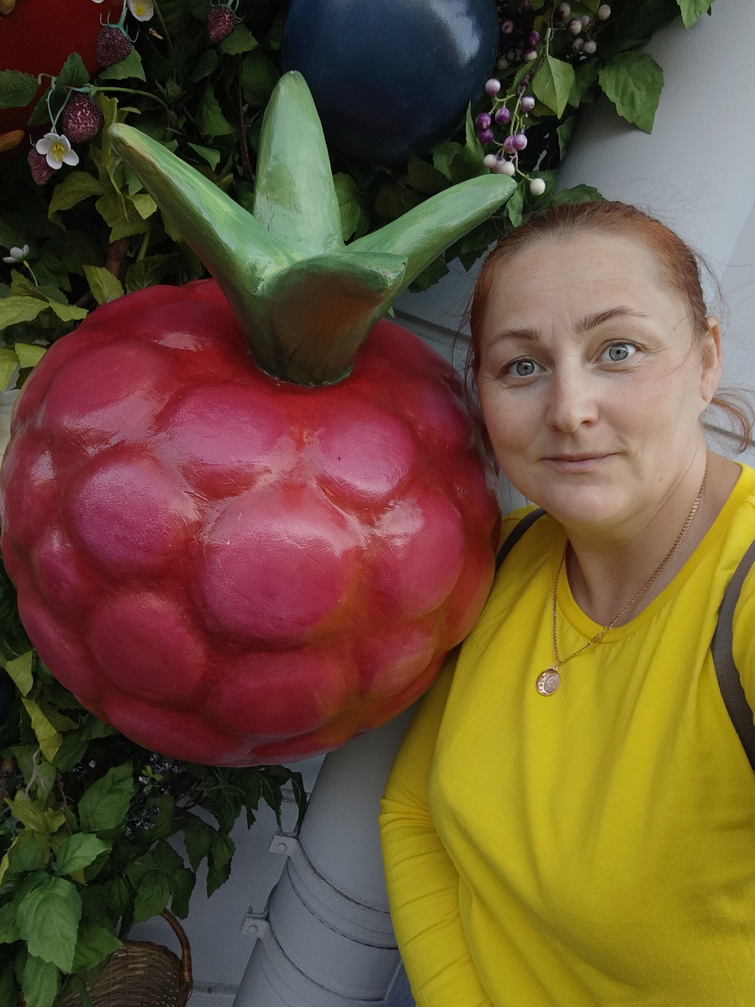 A woman with red hair and blue eyes poses for a selfie next to a giant, artificial raspberry. She is wearing a bright yellow long-sleeved shirt and a gold pendant necklace. Behind the raspberry, there are artificial green leaves and clusters of small purple and white berries, as well as a large dark blue sphere. The setting appears to be outdoors, with a light-colored, possibly painted, wall or structure in the background. The image was taken in the city of Saint Petersburg, Russia, likely in a park or decorative outdoor installation.