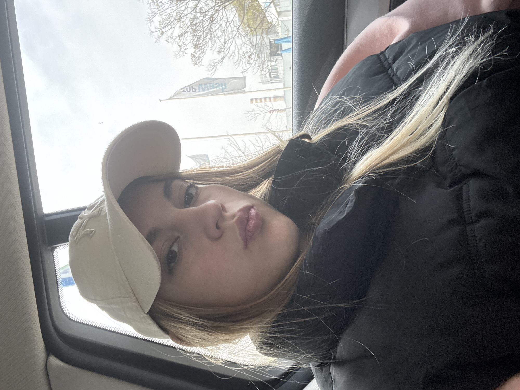 A young woman with fair skin and light brown hair with blonde highlights is wearing a light-colored baseball cap and a black puffer jacket. She is positioned inside a vehicle, near a window. Outside the window, blurred buildings and trees are visible, as well as a banner or flag with the text "ЛЕВЕЛЪТ". Based on the text on the banner, the location could potentially be Sofia, Bulgaria.