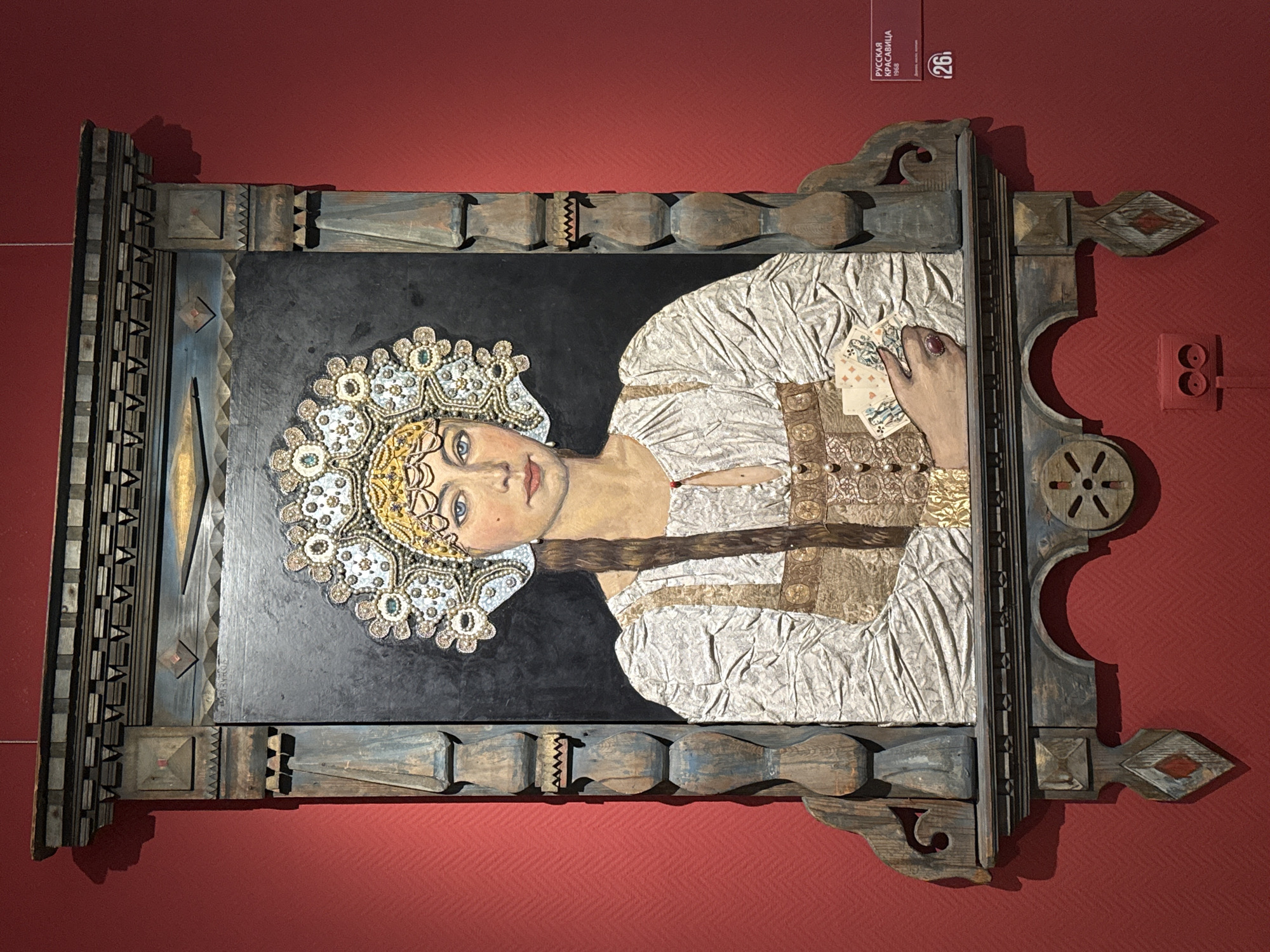 The image shows a painting of a young woman in traditional Russian attire, framed by an elaborately carved wooden border. The painting is displayed on a textured red wall, characteristic of museum settings. The woman is depicted with blue eyes, a light complexion, and a braid of brown hair. She wears a white blouse with intricate textured fabric and an ornate headdress adorned with jewels and pearls. In her hand, she holds a fan of playing cards. A small red label in the lower right corner of the image indicates the artwork is titled "РУССКАЯ КРАСАВИЦА" (Russian Beauty) and was created in 1968. Below this, a number "26" is visible. This artwork is located in the Novosibirsk State Art Museum in Novosibirsk, Russia.