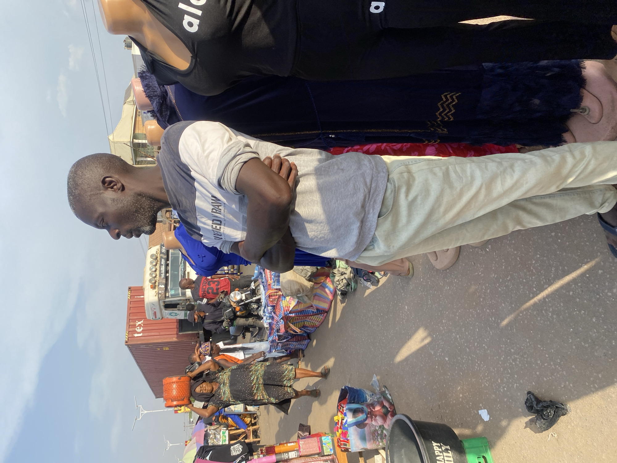 An outdoor market scene in Sabon Tasha, Nigeria, is depicted under a bright sky. In the foreground, a dark-skinned man with greying hair and a beard stands facing left with his arms crossed. He wears a grey and white long-sleeved shirt with "GED RAW" printed on it and light-colored pants. To his immediate right are two partially visible mannequins: one in a dark blue, fringed garment with gold patterns, and another in a black athletic top with "alo" text and matching black pants.
Behind the man, a paved street or market path extends into the midground. A woman in a patterned dark dress walks from left to right, balancing an orange gas cylinder on her head. To her right, a line of market stalls displays various goods, including colorful bags and fabrics, on the ground and low tables. Several other individuals are present: a man in a red "Chicago 23" shirt stands near a parked motorcycle, and another individual in a black cap and jacket is also visible.
Further back, a large white semi-truck with a red shipping container marked "tex" is positioned on the road. Overhead, utility wires traverse the sky. In the background, buildings with varied architectural styles are visible, including a light-colored structure with a pyramidal roof. The ground in the foreground shows shadows from objects outside the frame and contains a black plastic bucket on a green crate.
