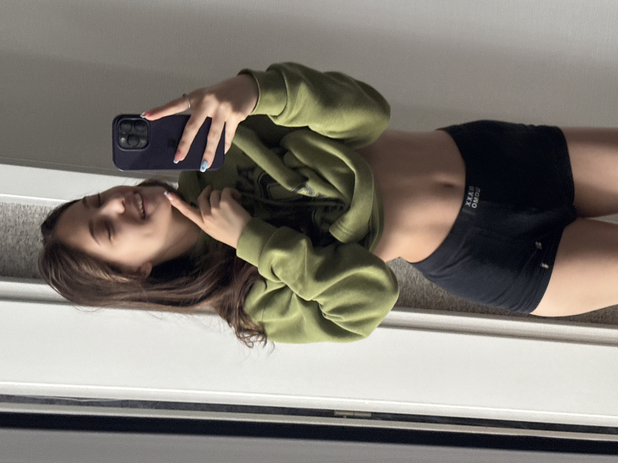 A young female is depicted taking a mirror selfie indoors, captured from a tilted perspective as if she is lying down. She has long, dark brown hair and a bright smile, with one hand holding a dark-colored smartphone (likely an iPhone) to take the photo and the other hand playfully pointing to her cheek. She wears a relaxed, oversized olive green hoodie, which is pulled up slightly, exposing her midriff, and black athletic short shorts. Her fingernails are painted with a light blue or swirled design, and she has a subtle ring on her left ring finger.
The setting appears to be a room with a plain light-colored wall and a light grey carpet, visible in the background reflection. The lighting is soft and even, suggesting daytime. On the waistband of her shorts, white text is visible, which appears to be "OMDU XXAW" when viewed in the mirror, likely reflecting a brand name like "MAXX DUOMO" or similar in reverse. The overall mood is casual and cheerful.