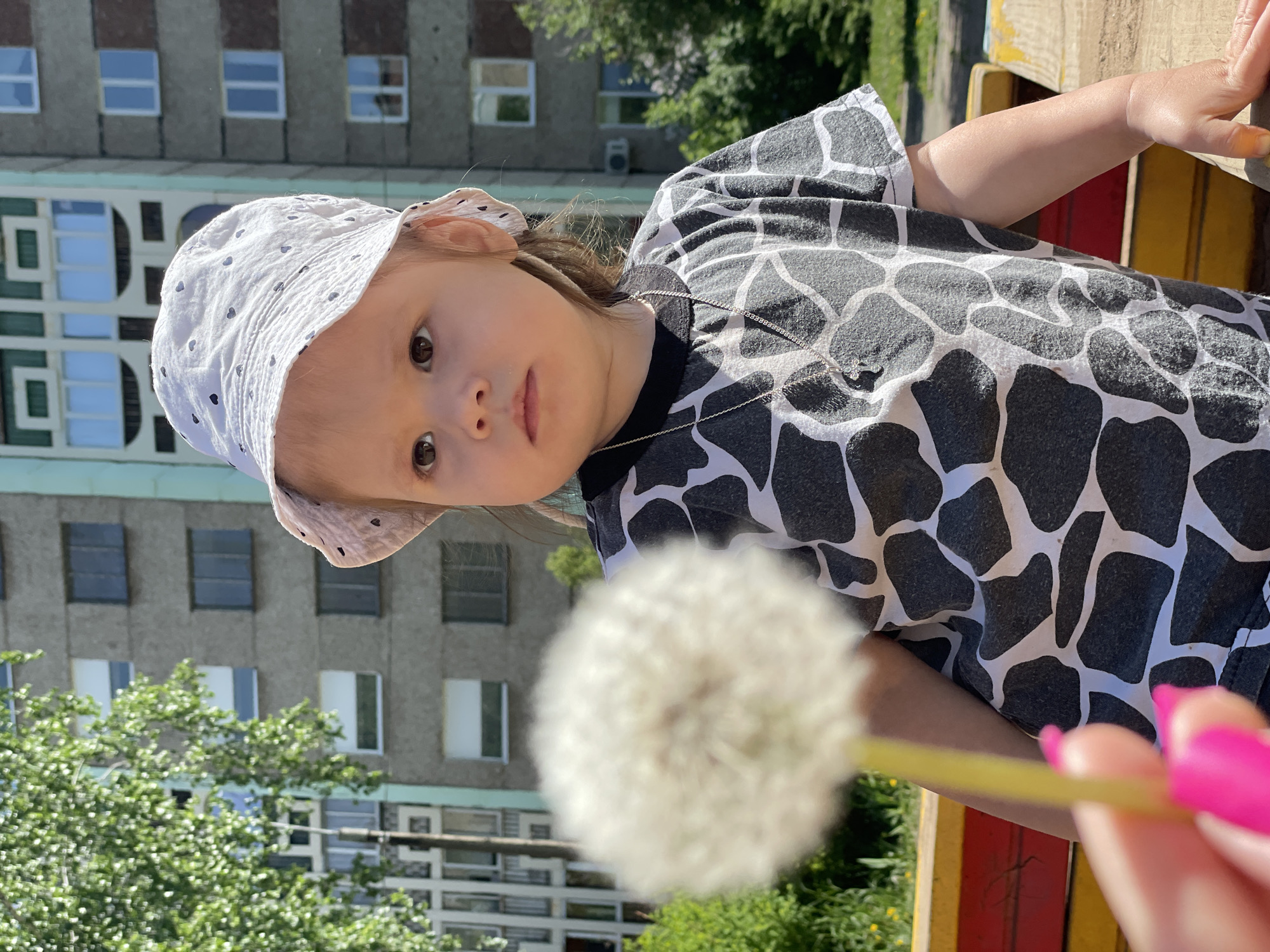 A young child wearing a white hat with polka dots and a black and white giraffe-print shirt is holding a dandelion. A building with multiple windows is in the background, along with trees. The location is Izhevsk, Russia. A hand with pink fingernails is holding the dandelion's stem. The child is looking at the camera.