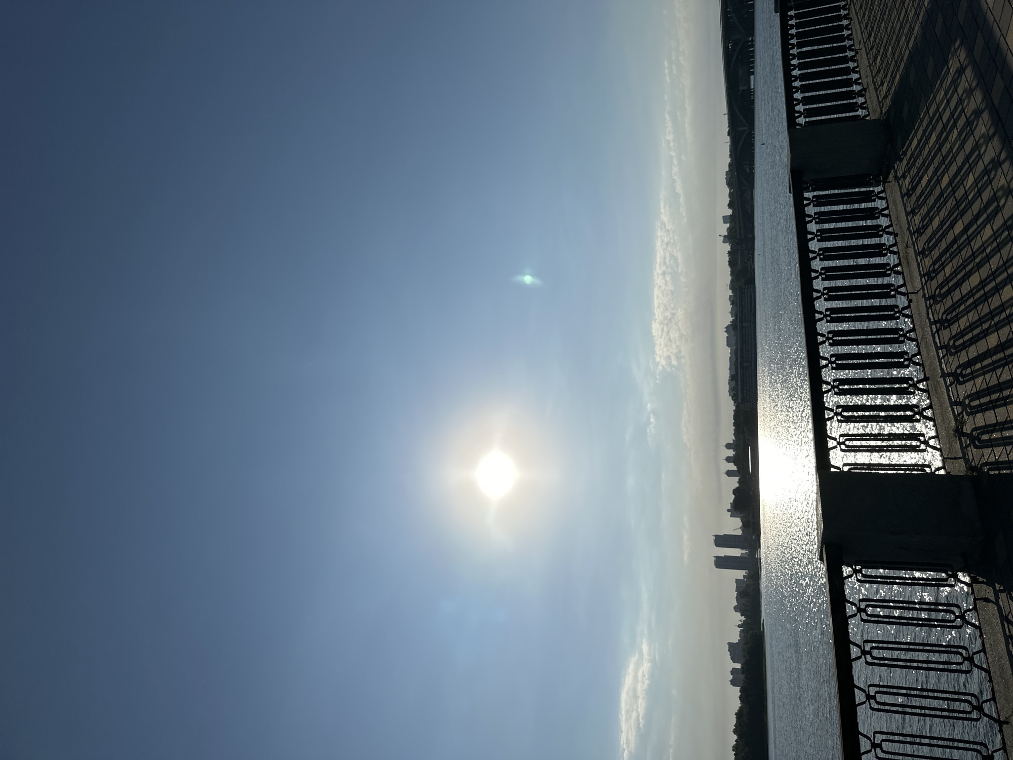 The image depicts a sunny day with the sun high in the sky, casting a bright glare on the water. In the foreground, there is a decorative metal fence and a paved walkway with long shadows, suggesting it's either morning or late afternoon. Beyond the walkway, a wide river flows, its surface shimmering with sunlight. On the opposite bank, a cityscape is visible, featuring a line of buildings and trees, with a bridge arching over the water in the distance. The sky is clear blue with a few wispy clouds. This scene is likely the embankment of the Dnipro River in Kyiv, Ukraine, given the architectural style of the buildings and the presence of the river and bridge, commonly seen along its banks. No people are visible in the image.