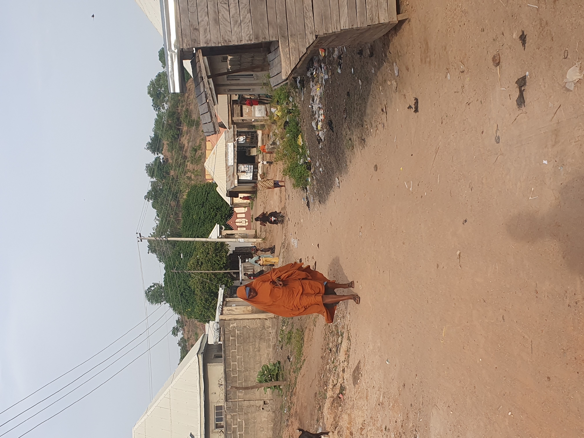 The image, rotated 90 degrees clockwise, depicts a street scene in a semi-urban or rural area under bright daylight, consistent with the provided context of Lokoja, Nigeria.
The setting is an unpaved, reddish-brown dirt road that appears well-trafficked and shows signs of debris. Various buildings line both sides of the road. On the right, a wooden structure, possibly a shop, stands next to a visible pile of refuse. Further along this side, more simple concrete block buildings are interspersed with vegetation. On the left, a building with a white metal roof and concrete block walls is prominent, with electrical poles and power lines running alongside it. Hills covered with dense greenery form the background.
The central subject is a person walking along the dirt road, wearing a long, flowing orange garment that covers most of their body, possibly barefoot or in sandals. Their head is turned slightly to the right, away from the direct camera view. Further down the road on the left, other people are visible, including one individual on a motorcycle and several others standing or congregating near buildings. A small, dark animal, possibly a dog, is partially visible in the lower-left foreground. The sky is clear, suggesting good weather.
Notable details include the widespread unpaved terrain, the mix of simple construction materials for the buildings, and the overhead utility lines. Faint and largely illegible text can be seen on a white sign on a building in the midground left, possibly starting with "GLOBAL" or "COMP", and some carved letters like "CA" or "GA" on the wooden building on the right. The scene captures a moment of daily activity in the locale.